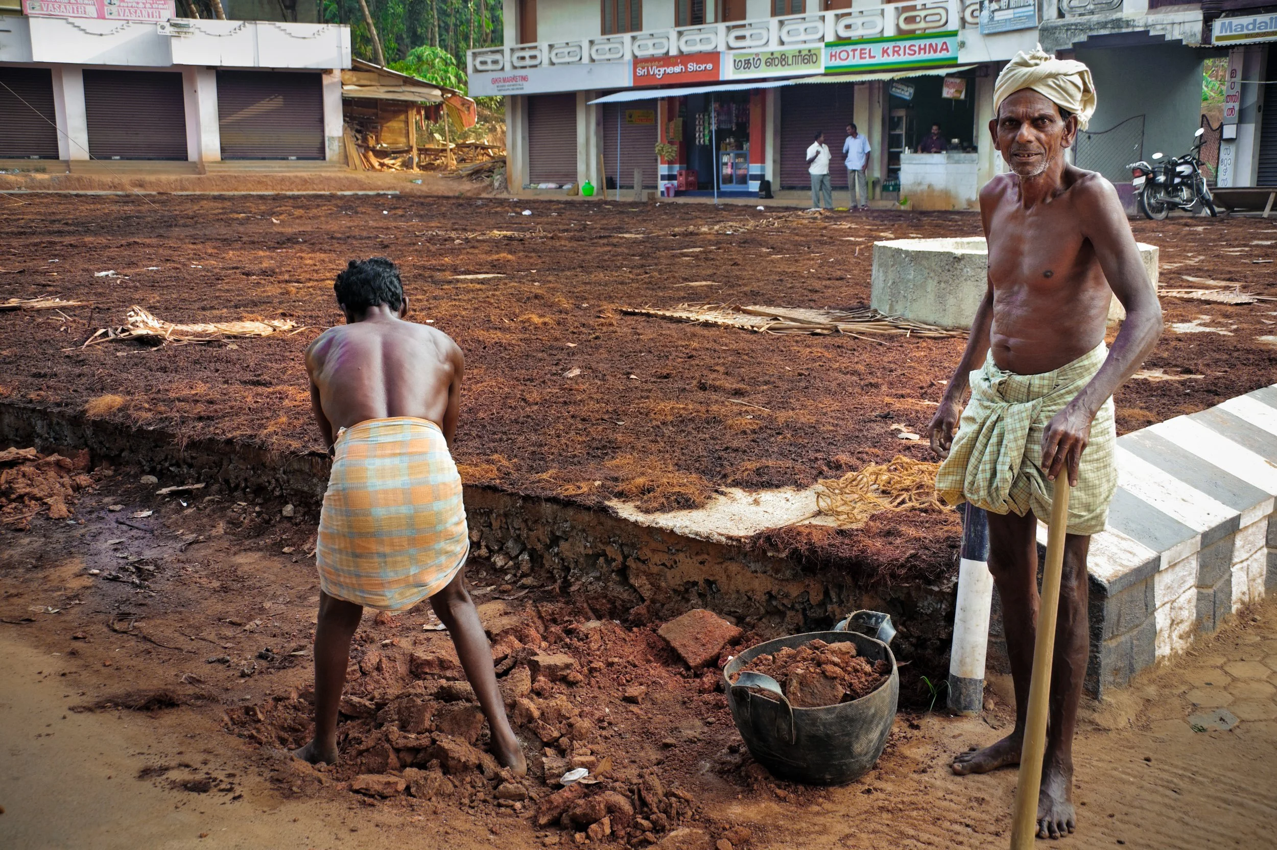 Two men working on a construction site in an urban area. One man stands holding a shovel, wearing a traditional dhoti and a turban, with a bucket of soil nearby. The other man, shirtless and wearing a similar cloth, is bent over working on the ground