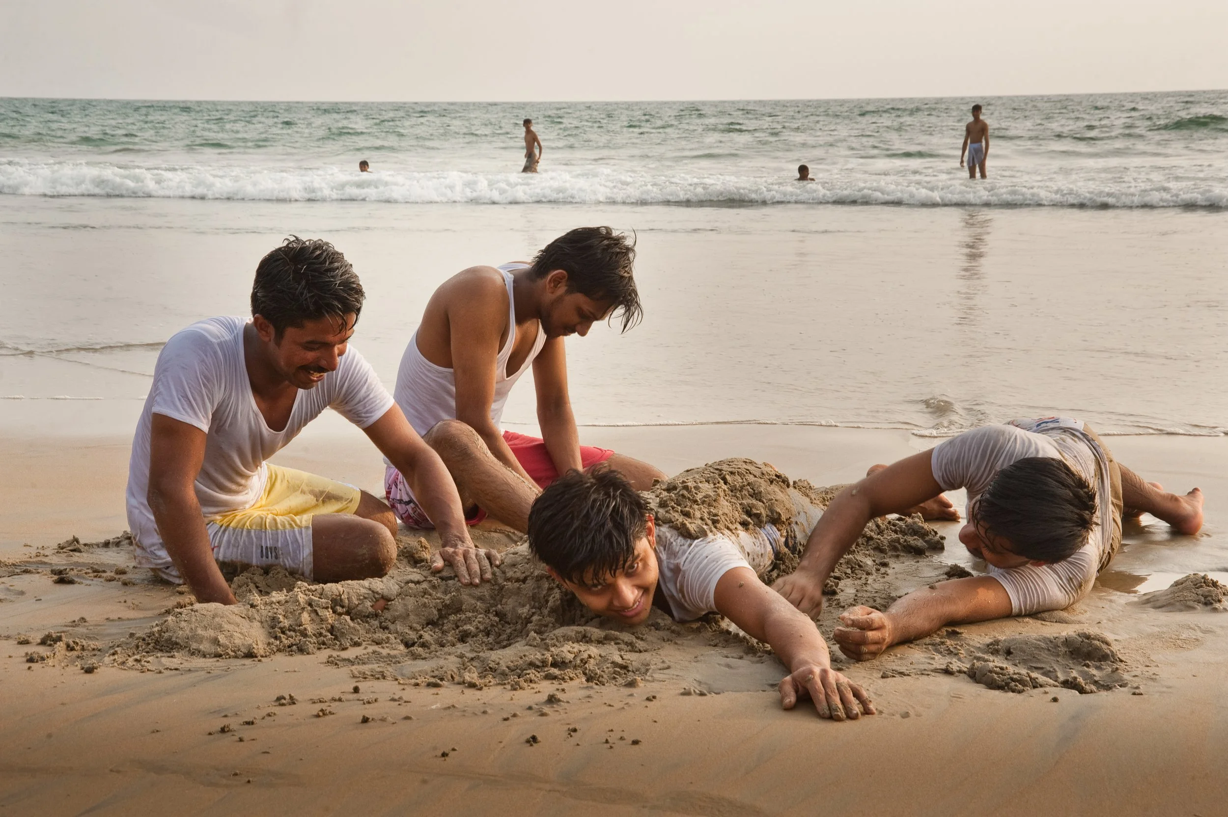 Four boys playing in the sand on the beach, with the ocean and other children in the background.