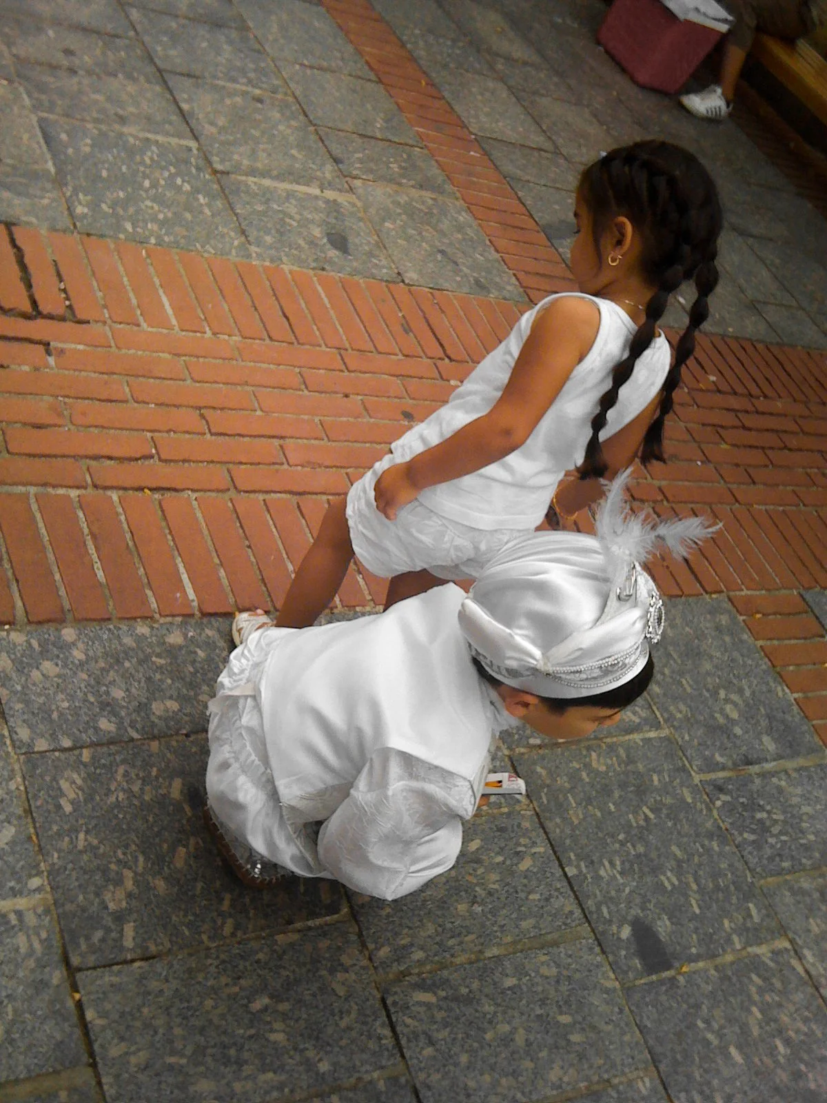 Two young children dressed in white outfits, one girl with braided hair standing and one boy with a white head covering kneeling on a tiled floor, with a background of a brick and stone pattern.