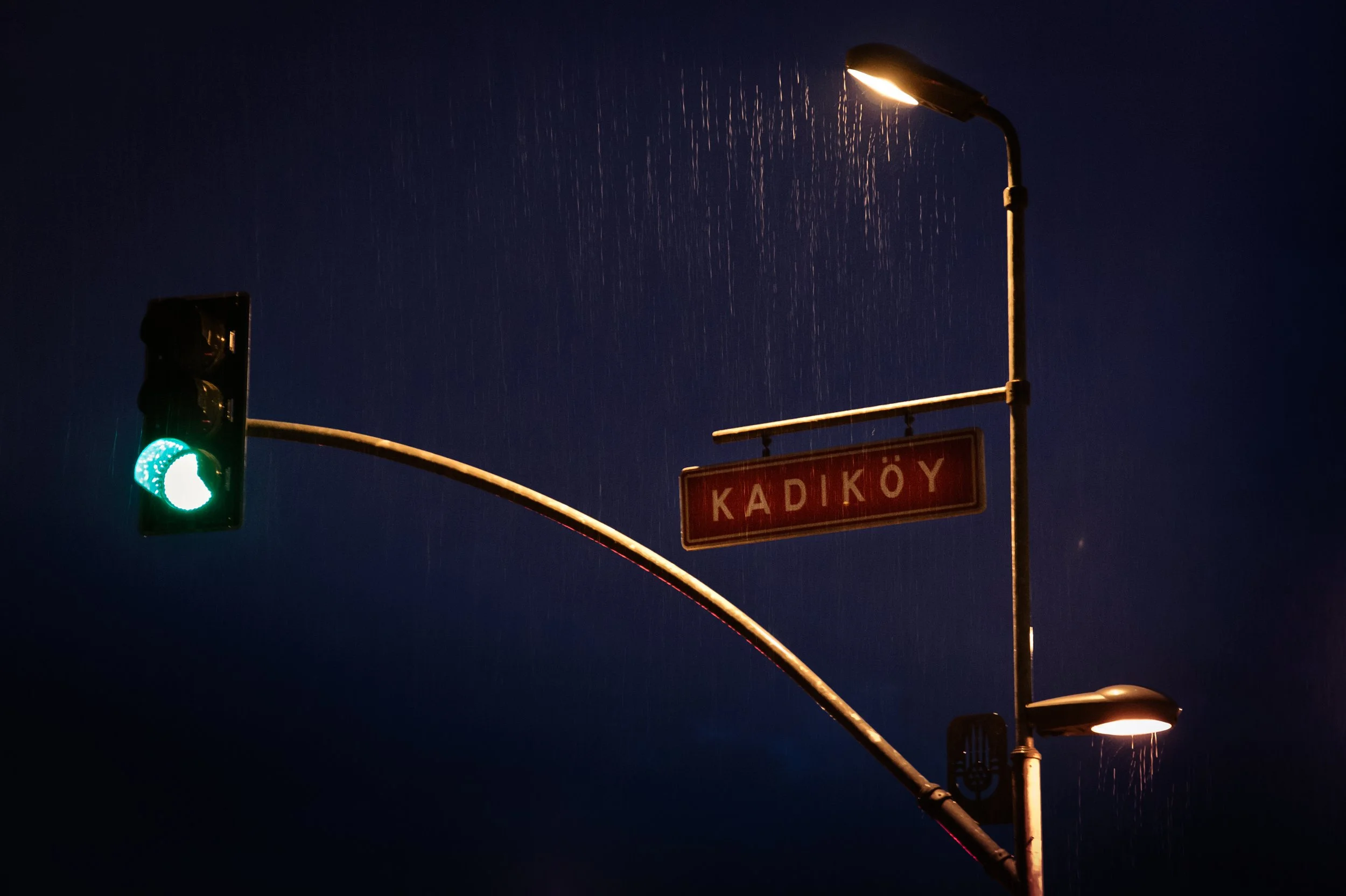 Nighttime traffic light showing green, street sign reading 'KADIKÖY', street lamps, and rain.