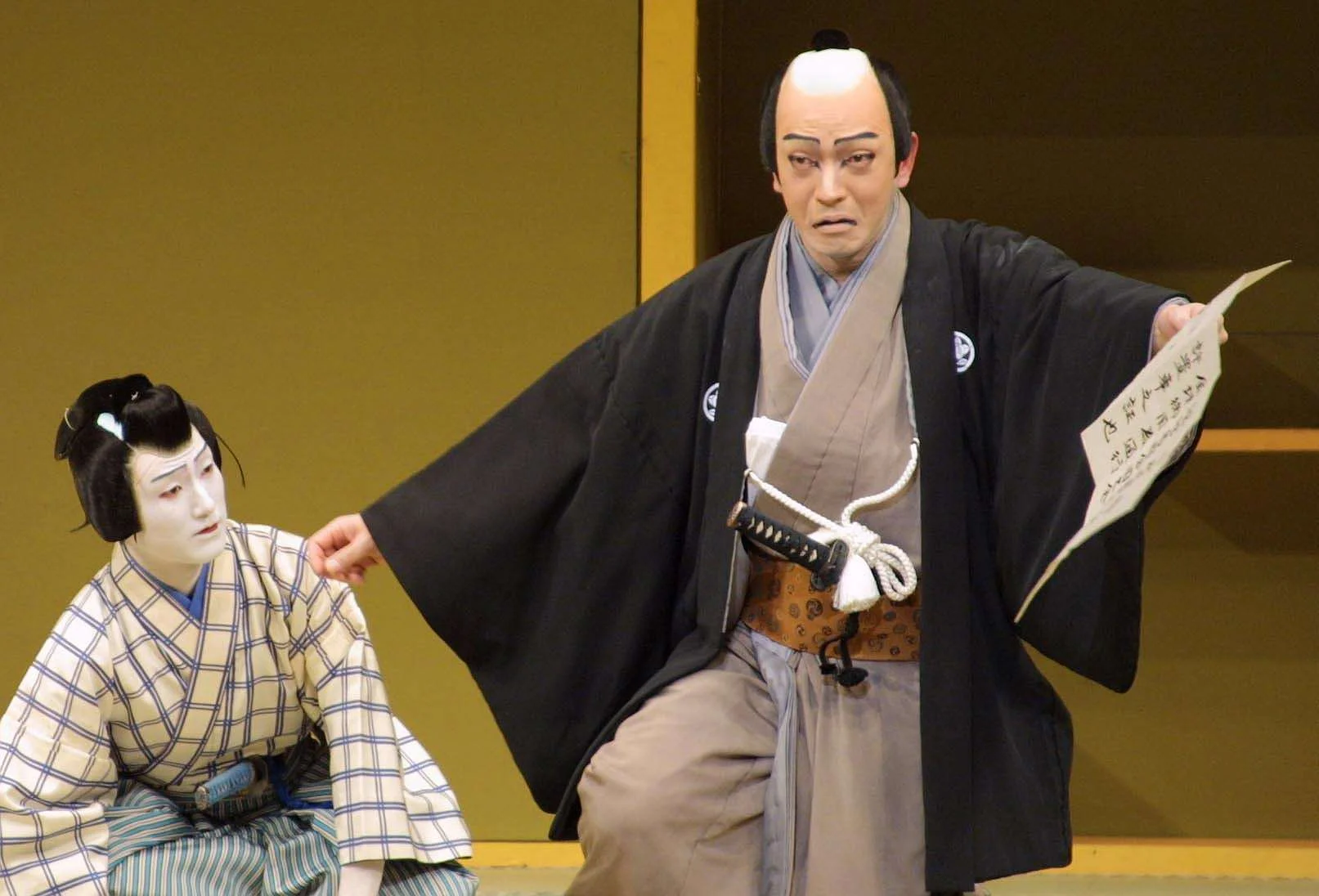 A traditional Japanese puppet show scene with two puppets dressed in historical clothing. One puppet appears as a man with a worried expression, wearing a black kimono and holding a script, while the other puppet is dressed as a woman in a kimono, wi
