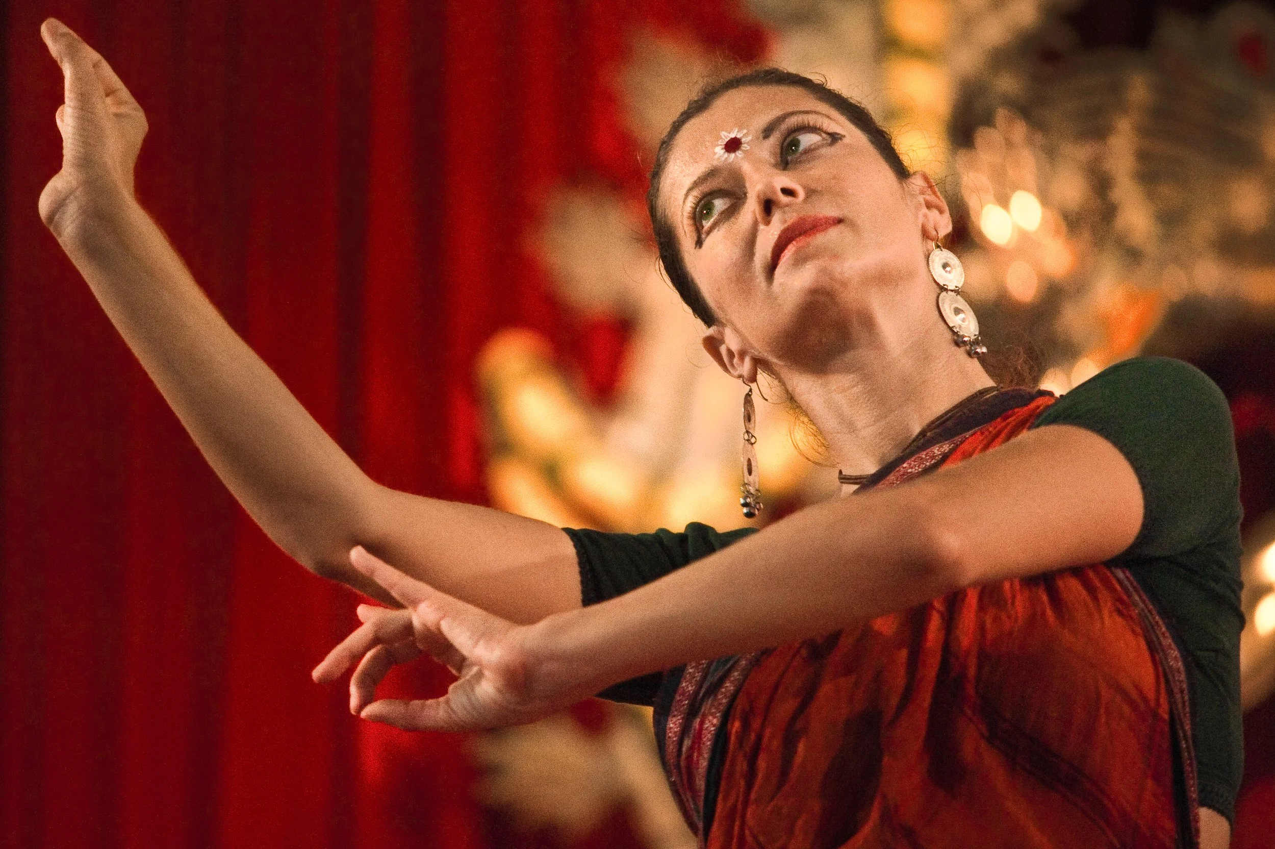 A woman performing a dance on a stage with a red curtain in the background, wearing earrings and traditional clothing, with her arms extended gracefully.