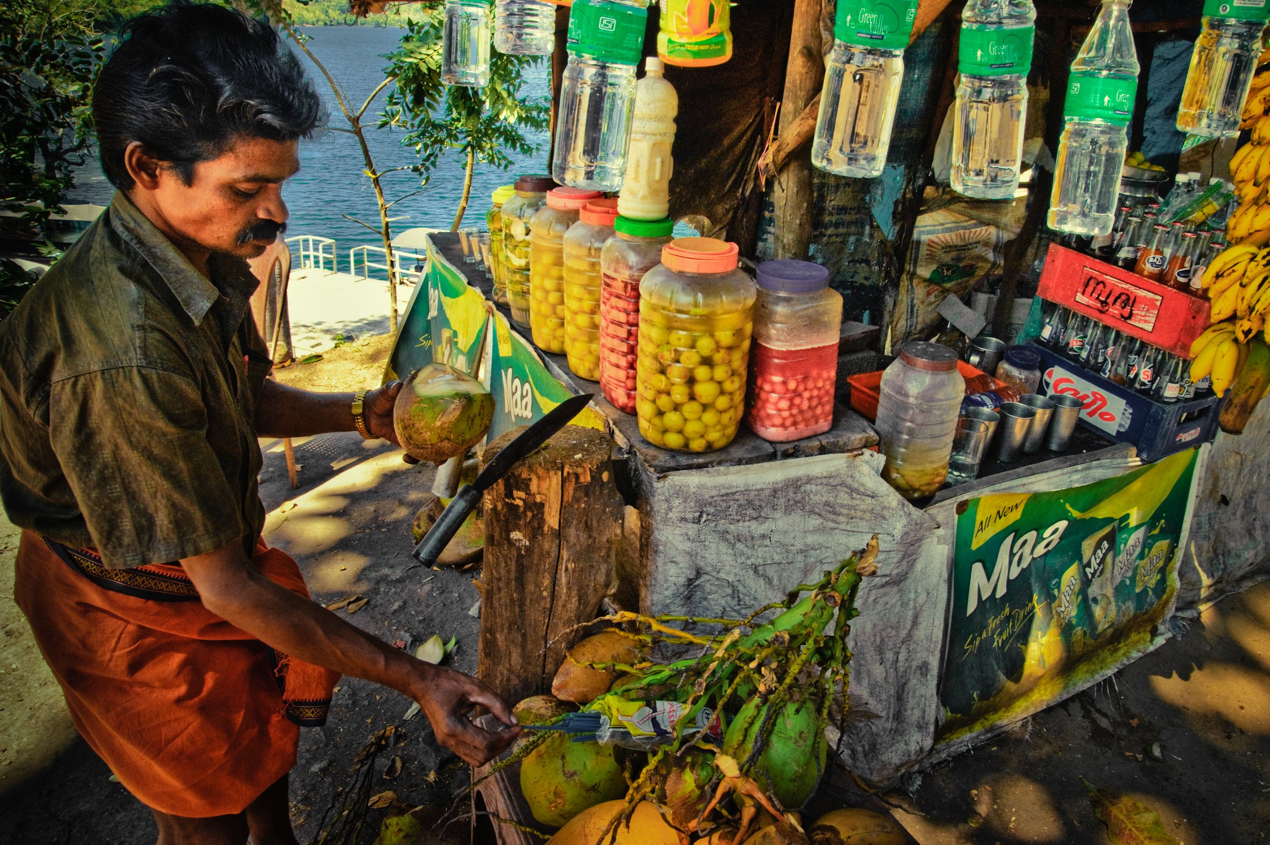 Man selling fresh coconuts, jars of sliced fruits, and snacks at a roadside stand near a lake or river, with bananas hanging from the stand.