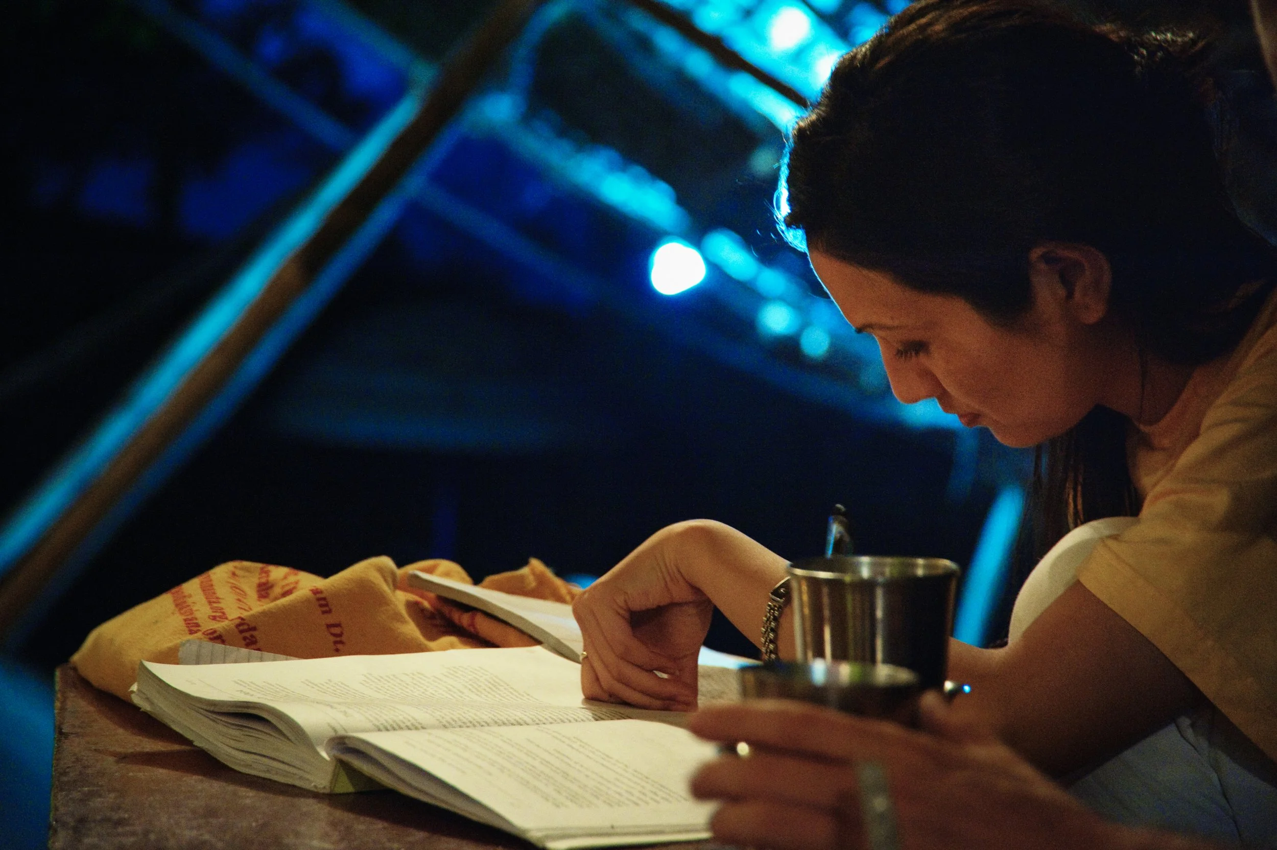 A woman reading a book at a table, with a metal cup in hand, under blue lighting in a dark environment.
