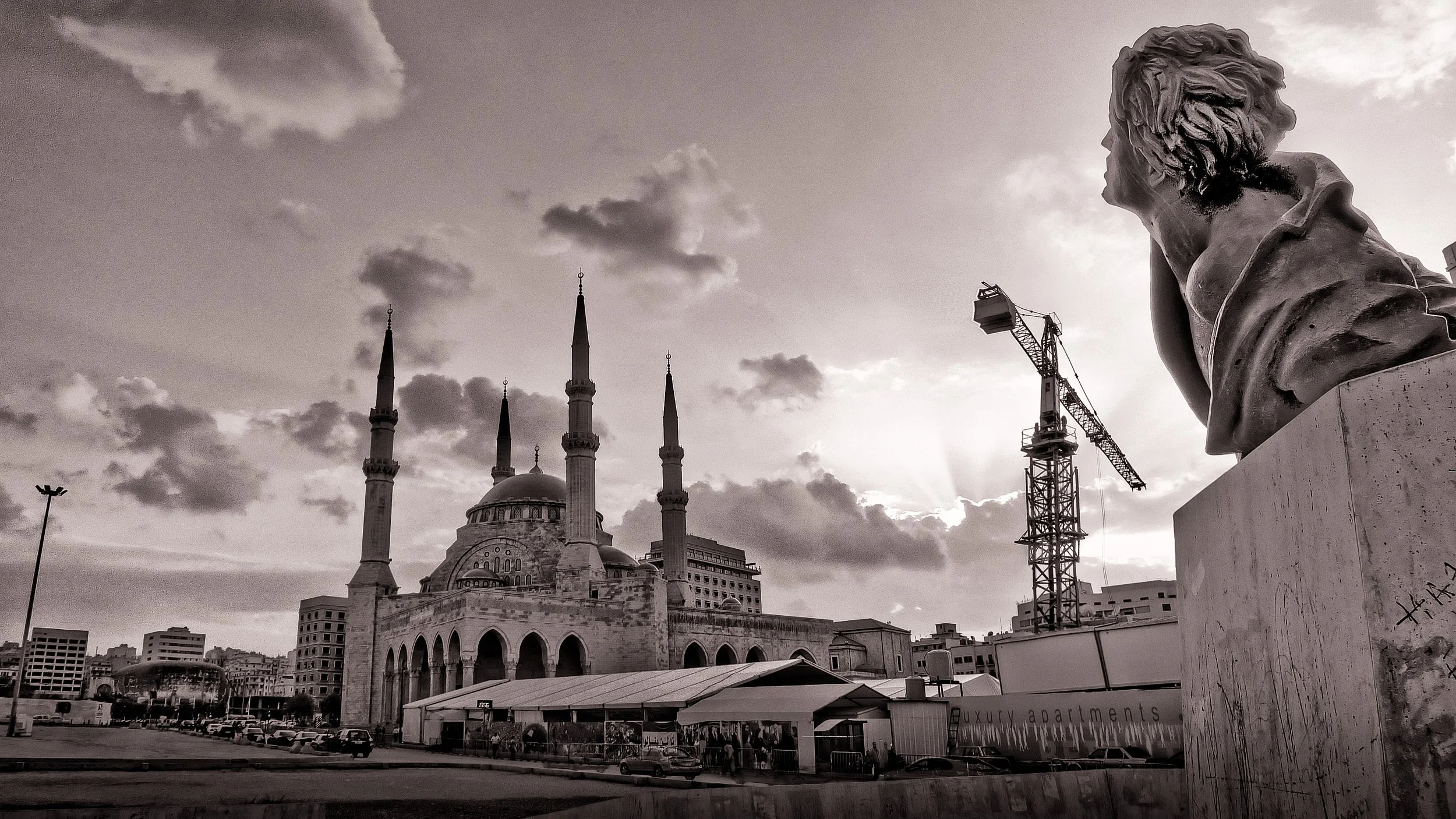 A black-and-white photo of a cityscape featuring a mosque with four minarets, a large dome, and a statue bust in the foreground to the right. There is a construction crane and modern buildings in the background.