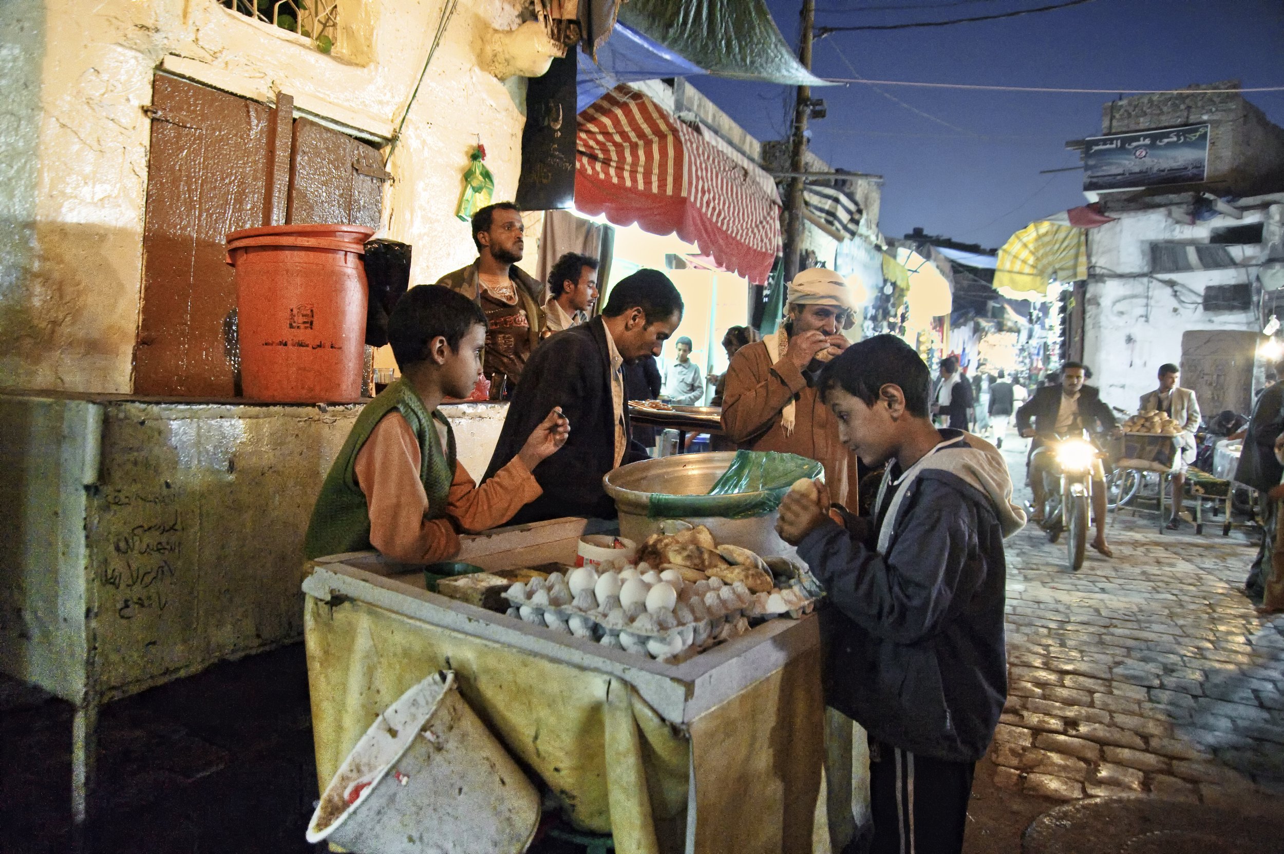 A busy street market scene at dusk with vendors and shoppers. A young boy in a jacket is purchasing eggs from a street vendor's stand. The stand displays eggs and other goods. People are walking and riding motorbikes in the background.