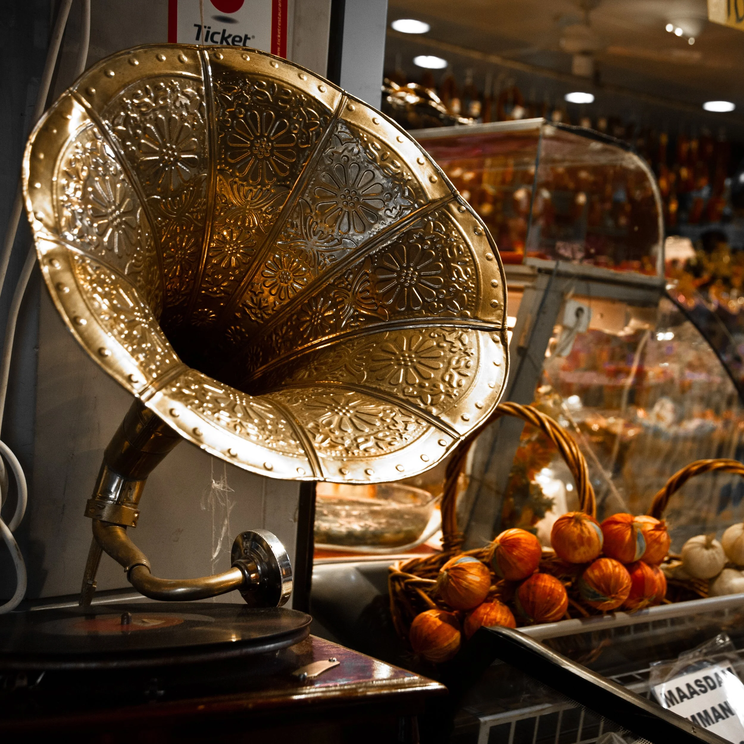 An antique brass phonograph with ornate floral patterns on the horn. Behind it, there are baskets of produce including onions and garlic, with a glass display case and colorful items in the background.