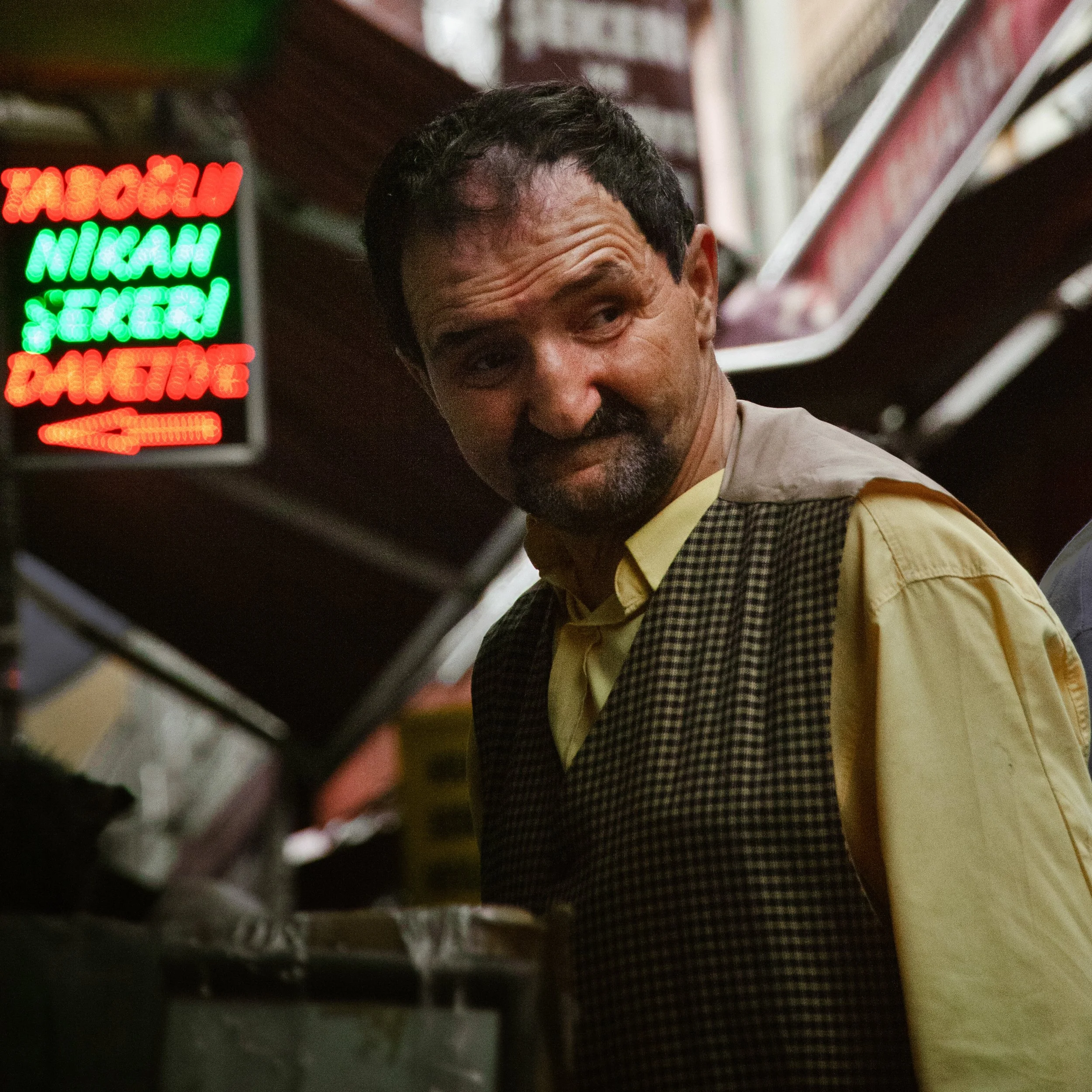 A man with dark hair and a beard, wearing a yellow shirt and checkered vest, looking to his left inside a store or shop with neon signs in the background.