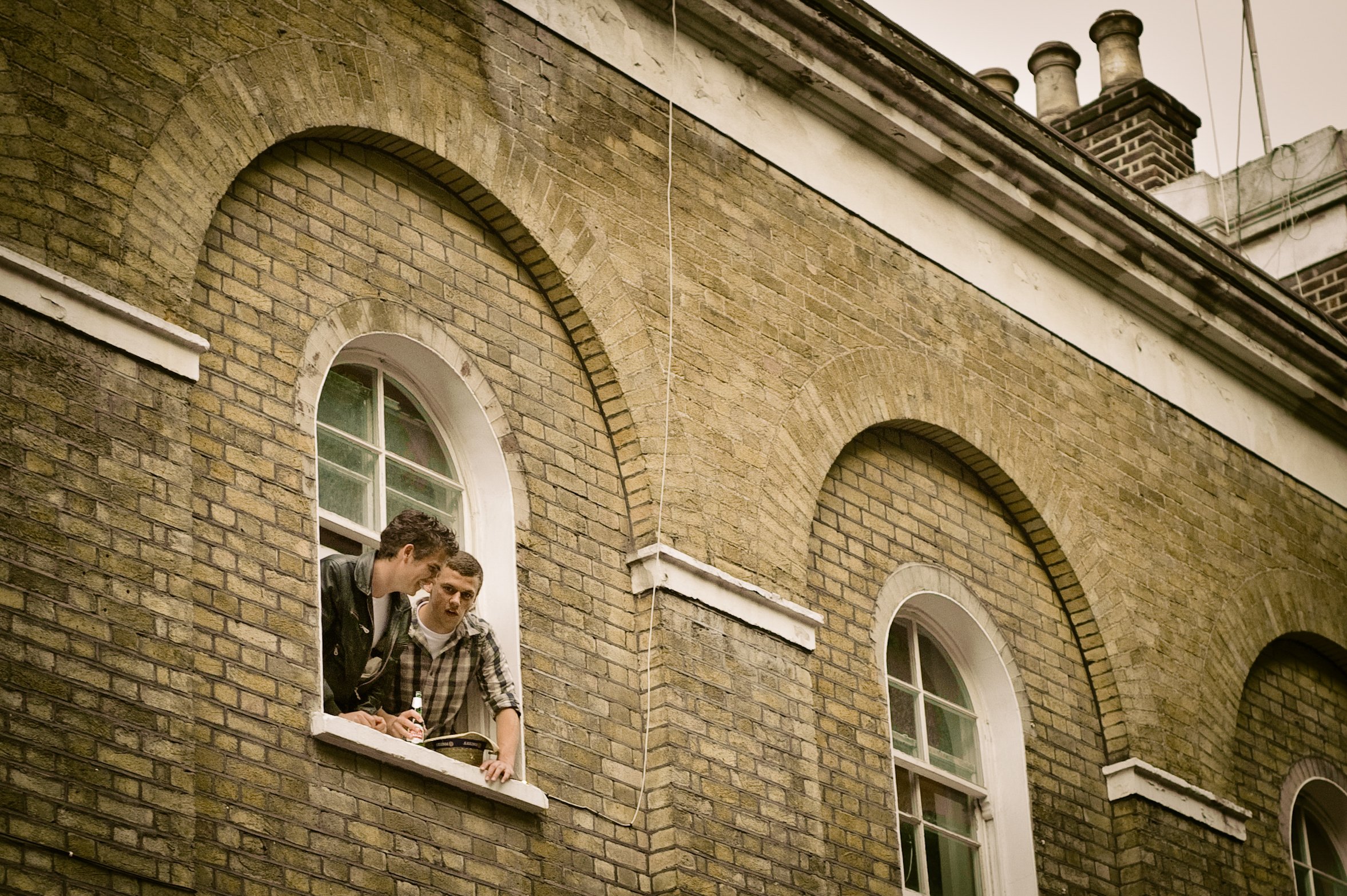 Two men leaning out of a window on the second floor of a brick building, engaged in a conversation, with one holding a screwdriver and the other with a phone and notebook, surrounded by arched windows and brick architecture.