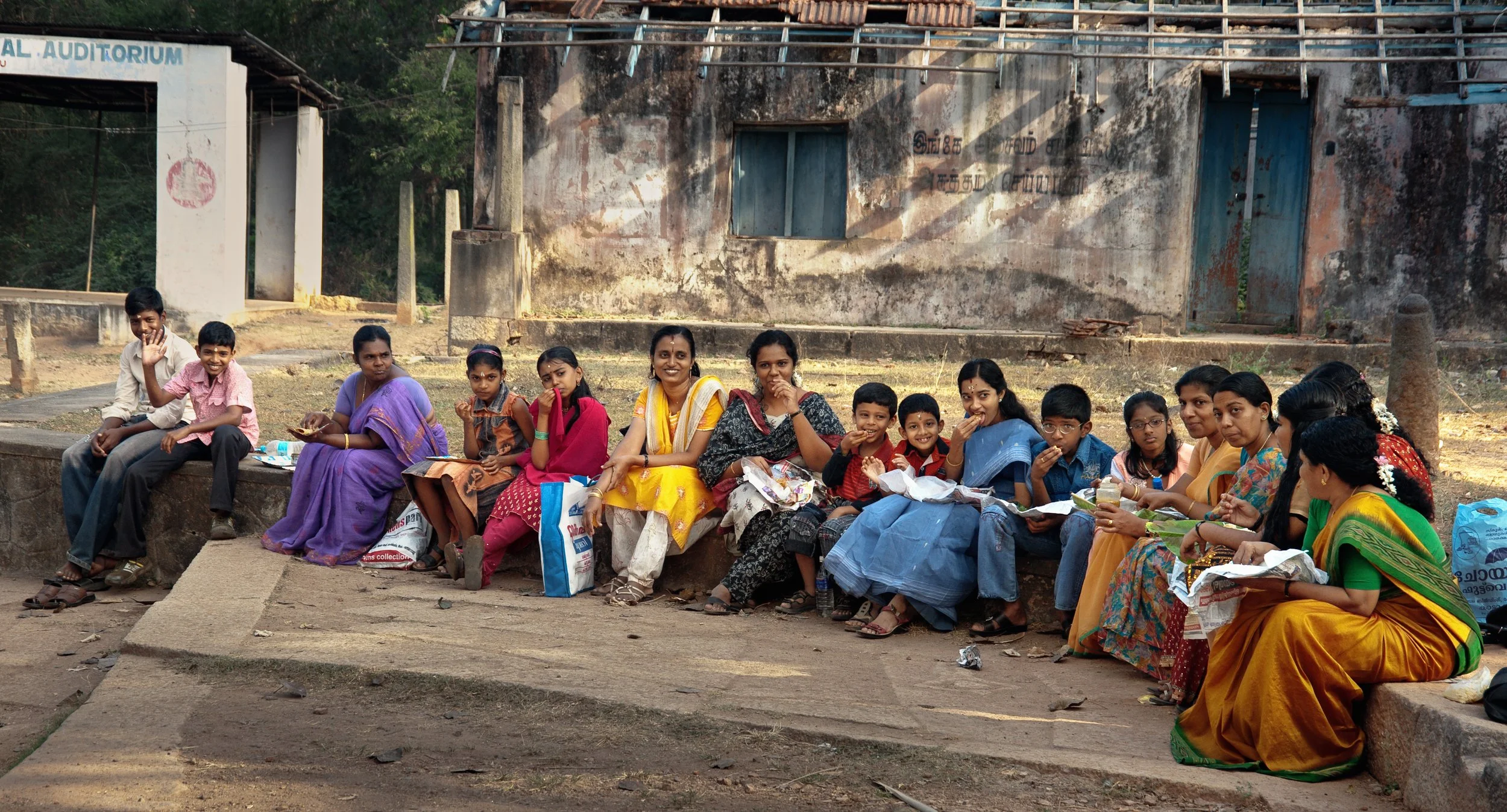 A group of children and women sitting on a low wall outside, smiling and eating together, with a weathered building and trees in the background.