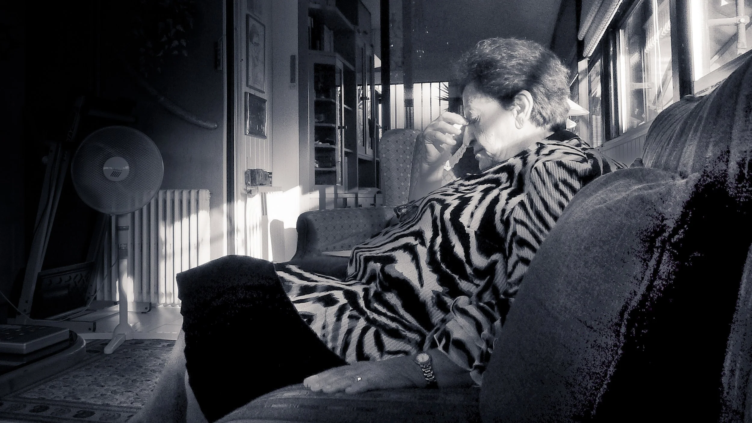 A woman with short hair sitting on a couch in a living room, wearing a zebra-print robe, touching her face, with sunlight streaming through the windows.
