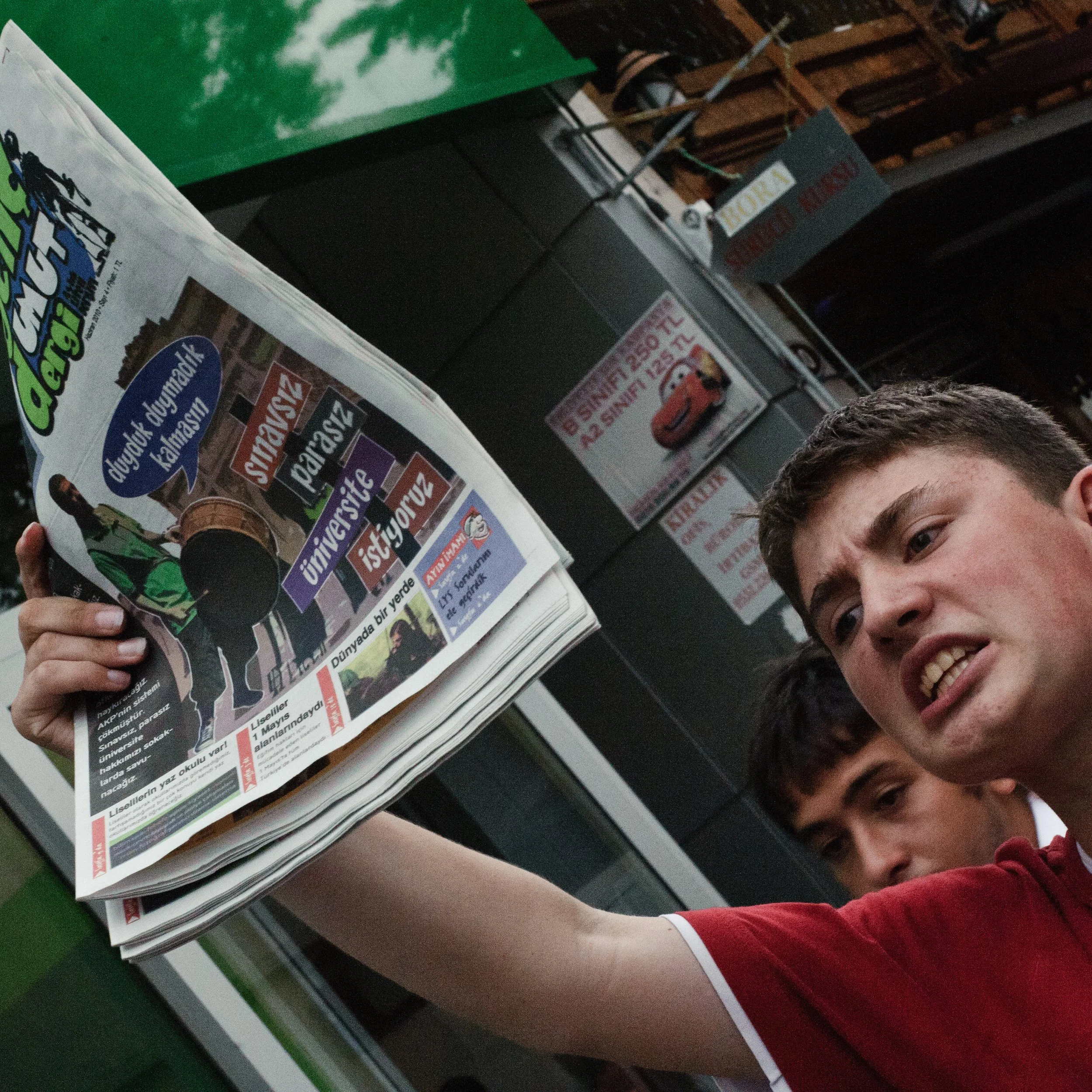 Young man with short hair and red shirt holding a newspaper, with other young men in the background, standing outside a building.