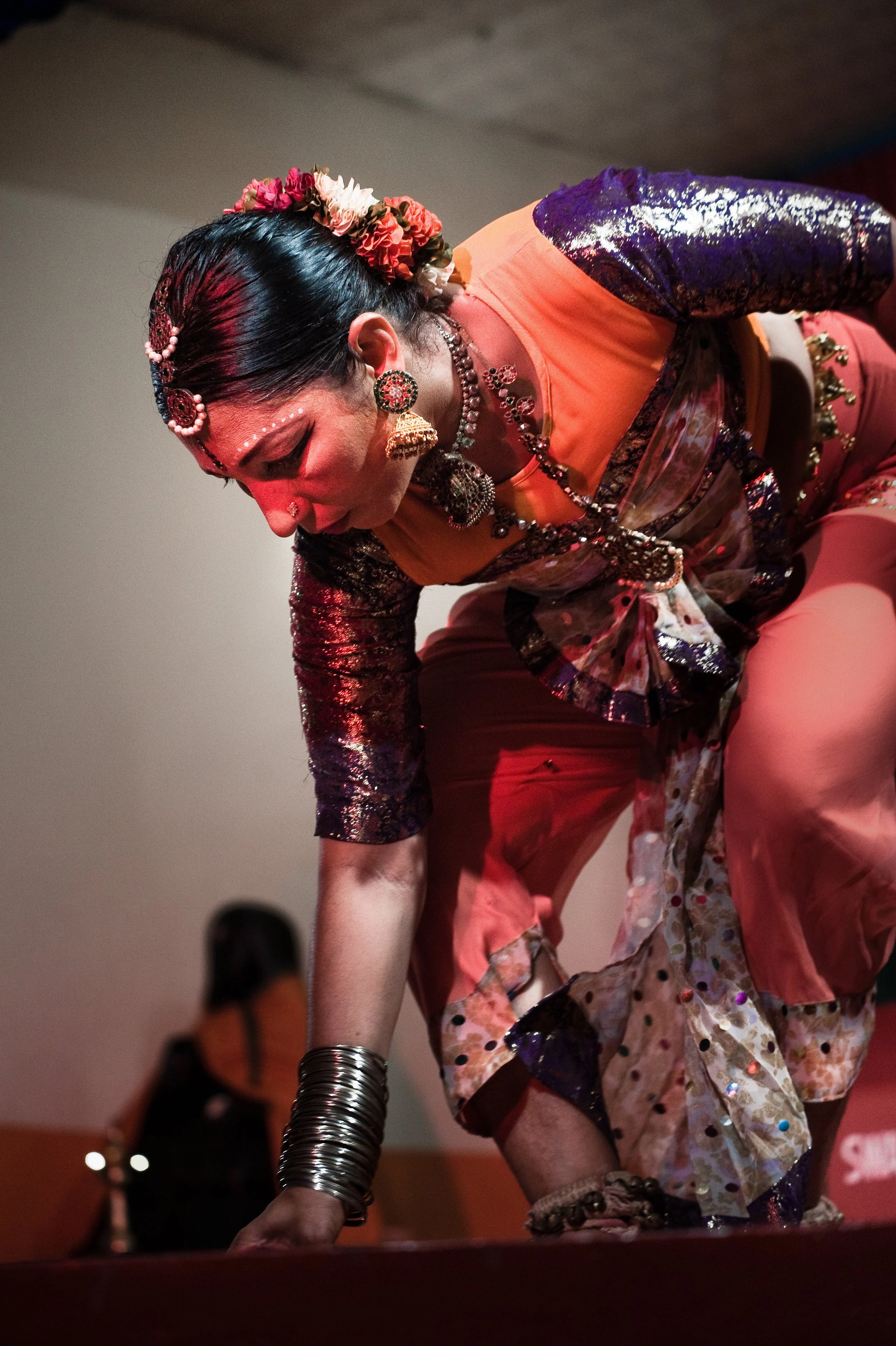 A woman dressed in colorful traditional Indian attire decorated with jewelry and flower accessories, performing a dance on stage.