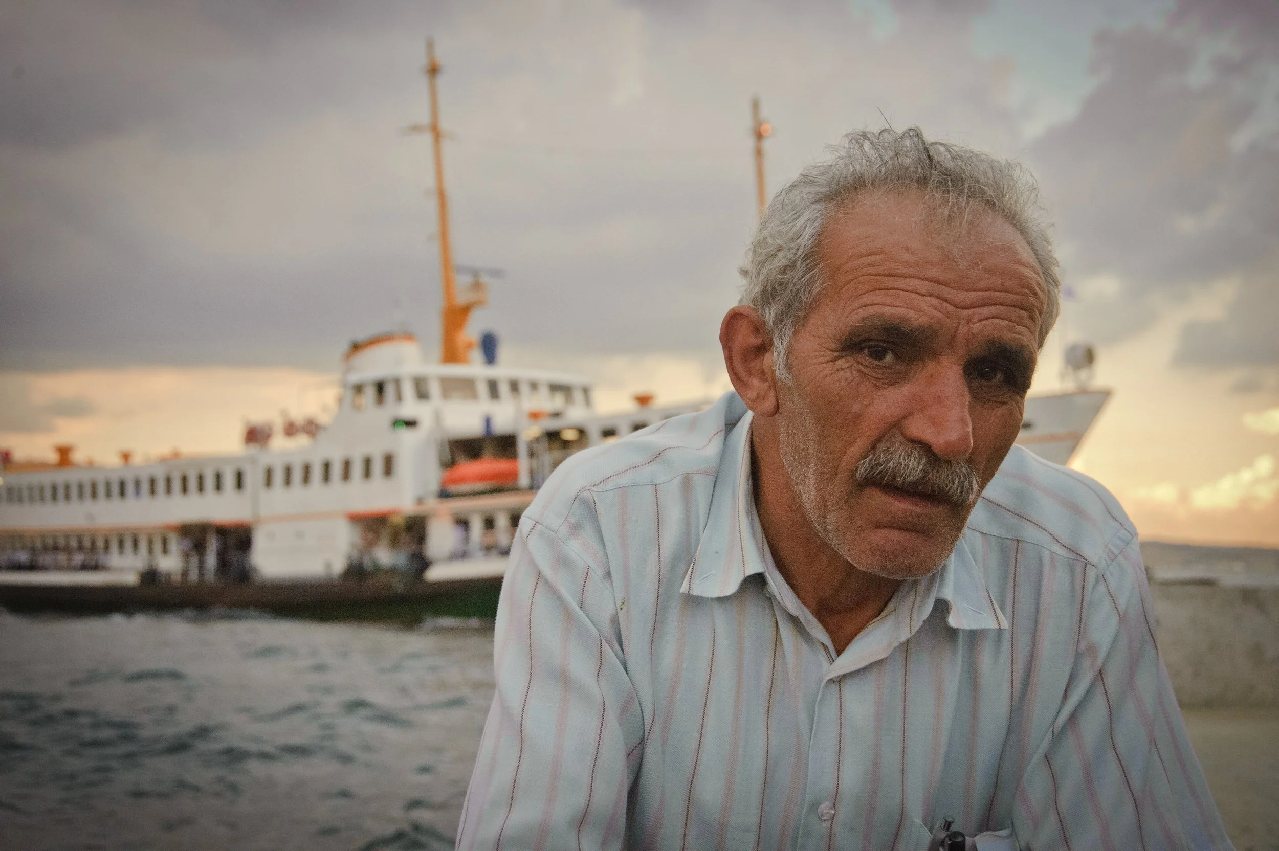 An elderly man with gray hair and a mustache wearing a striped shirt, looking at the camera with a large boat or ship in the background during sunset or sunrise on a cloudy day.