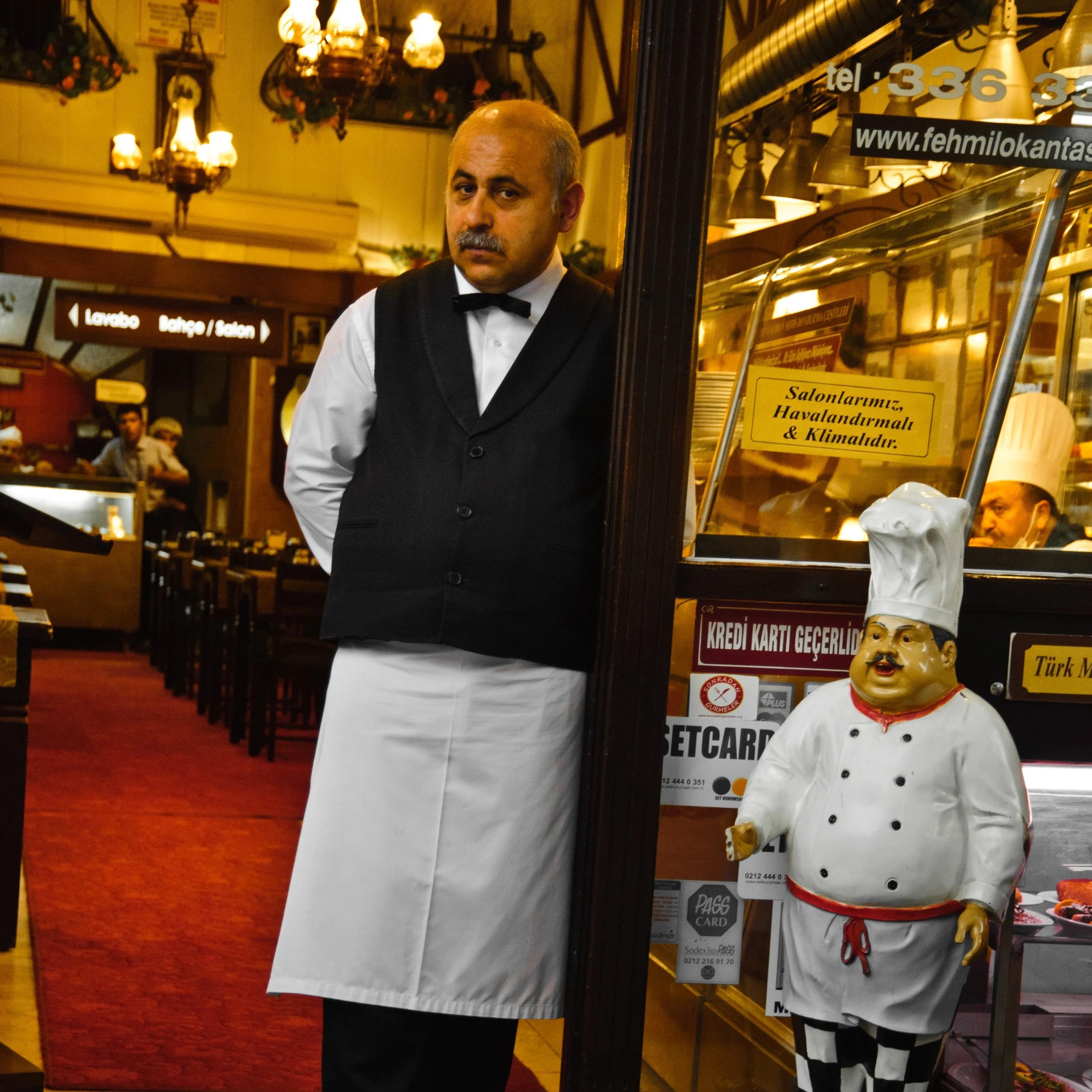 A waiter in a tuxedo with a white apron stands inside a restaurant, with a chef statue wearing a chef's hat and checkered pants outside the entrance.
