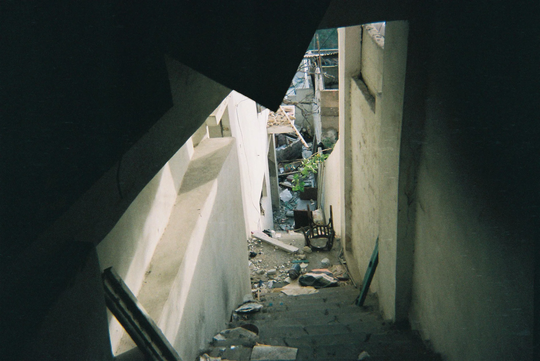 A narrow, steep staircase in a building leading down to a cluttered, dirty basement or lower level with debris, scattered objects, and a small plant.