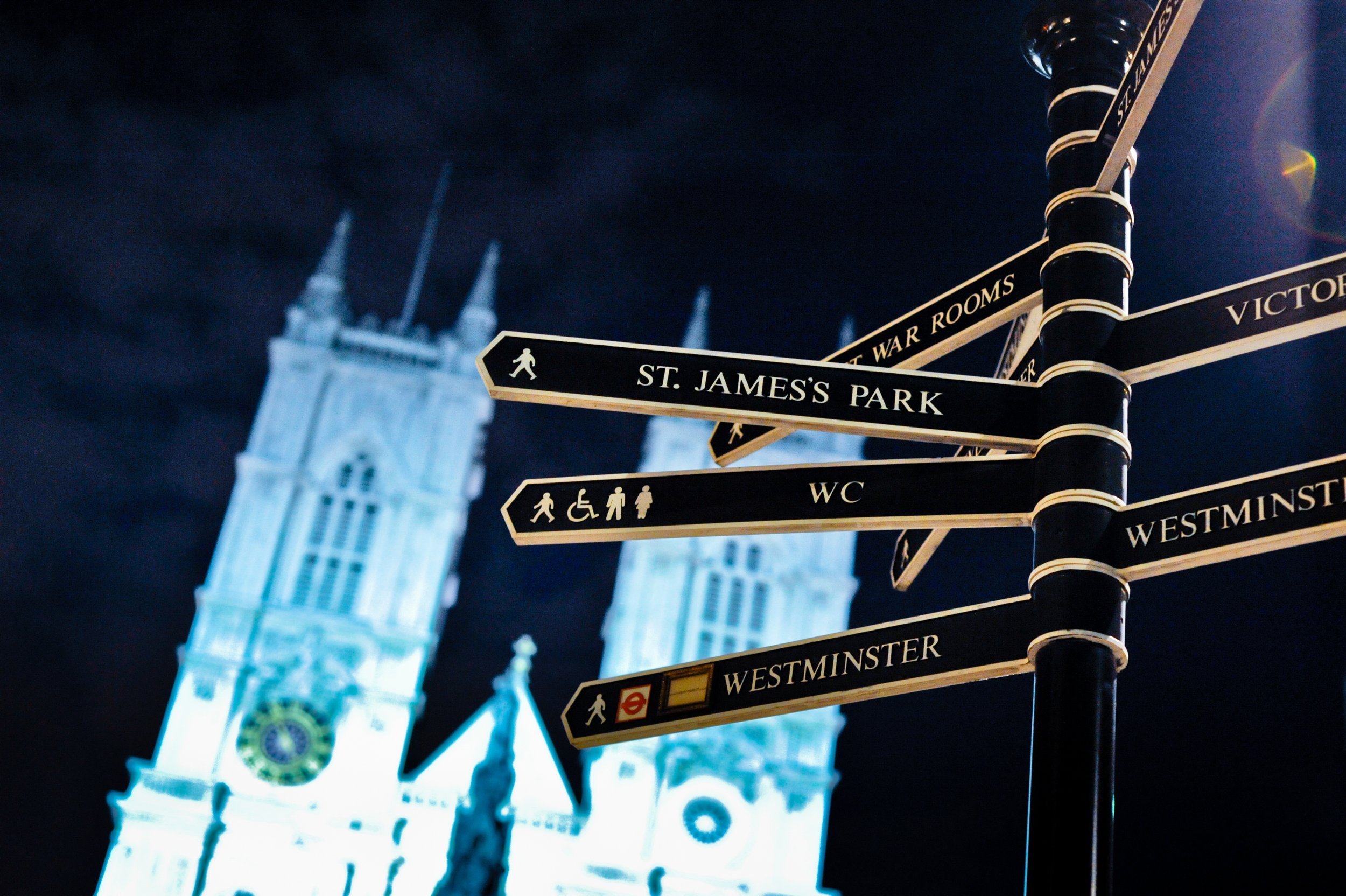 Signpost with directions to various locations in London, including St. James's Park, Westminster, and WC in front of Big Ben at night.