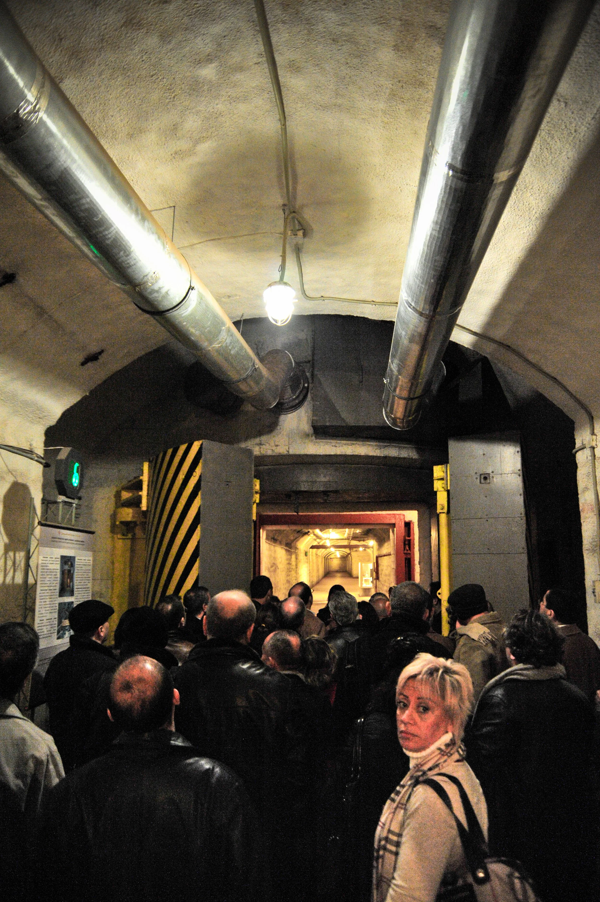 Crowd of people in a tunnel, possibly an entrance to a historical site or underground train station, with industrial pipes and lighting overhead.