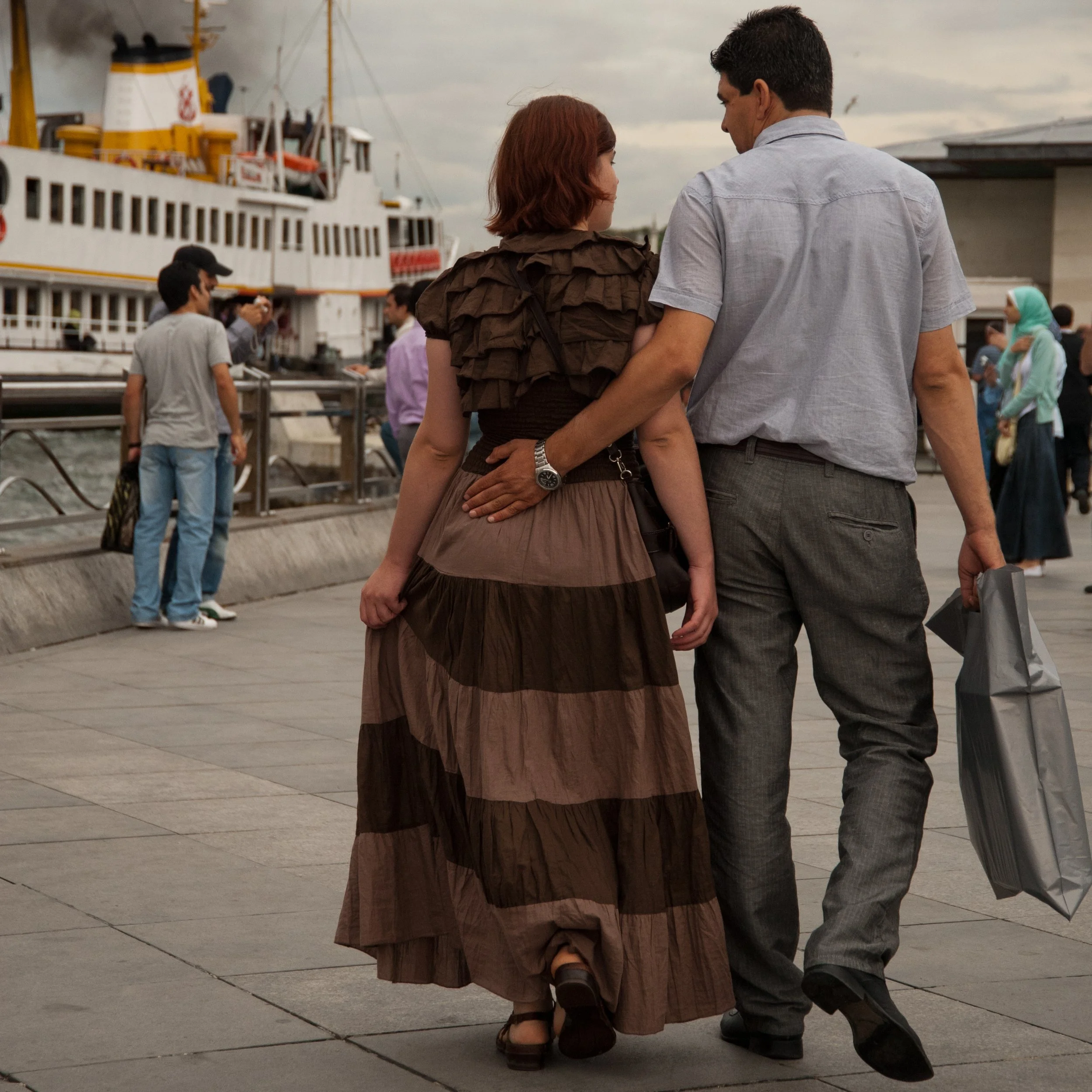 A man and woman walking together near a waterfront, with ships and other people in the background during a cloudy day.