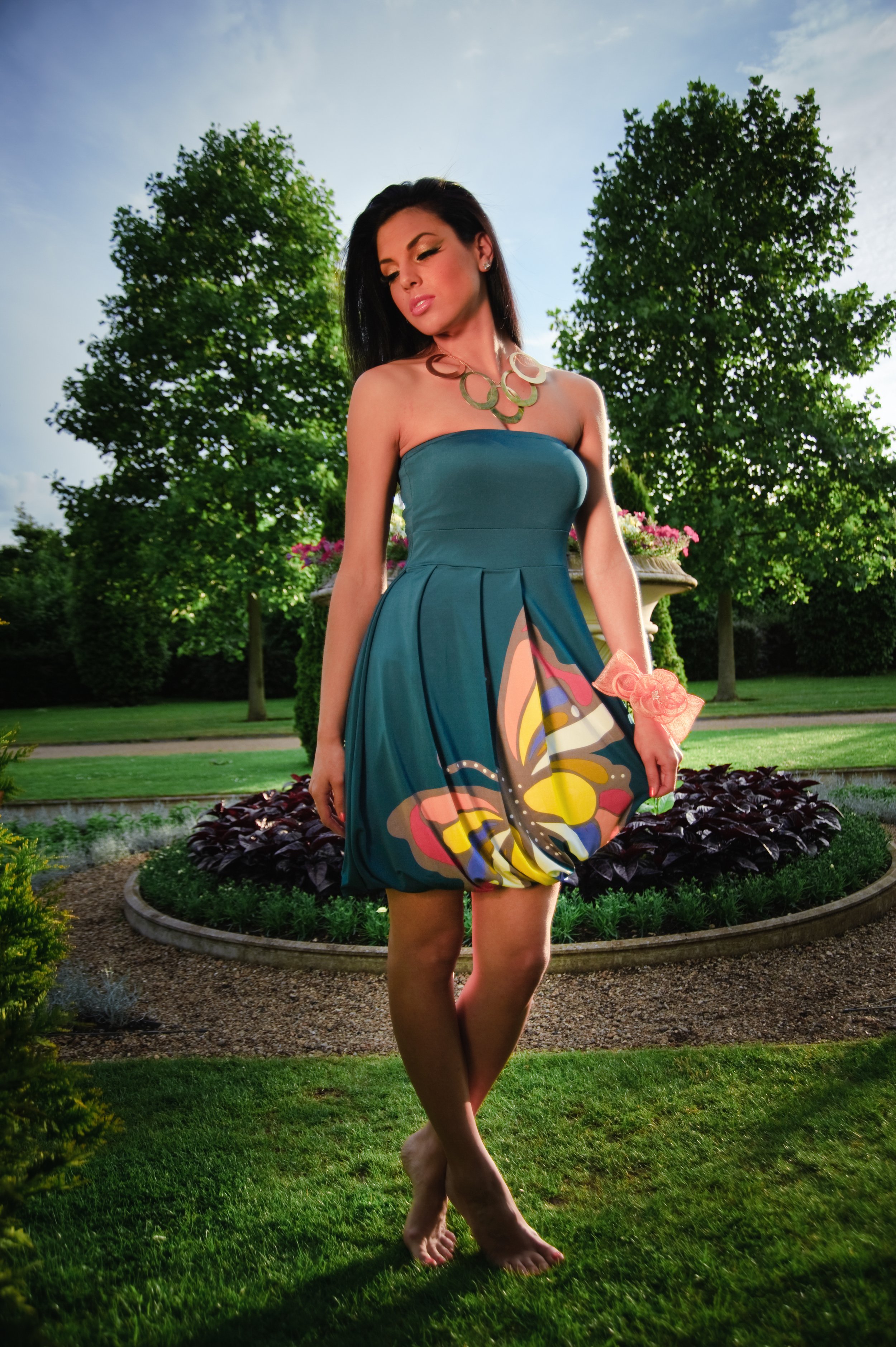 A woman standing barefoot in a garden wearing a strapless teal dress with a butterfly print on the skirt, a large necklace, and a pink ribbon bracelet, with trees and a lawn in the background.