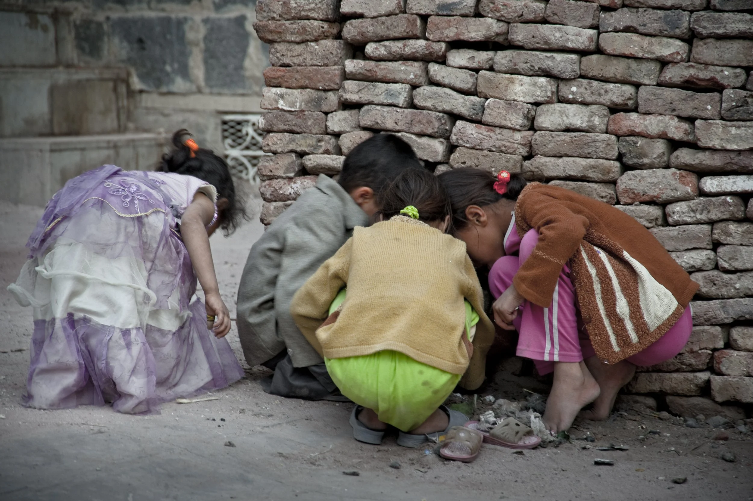 Four children crouch near a brick wall, looking at something on the ground.