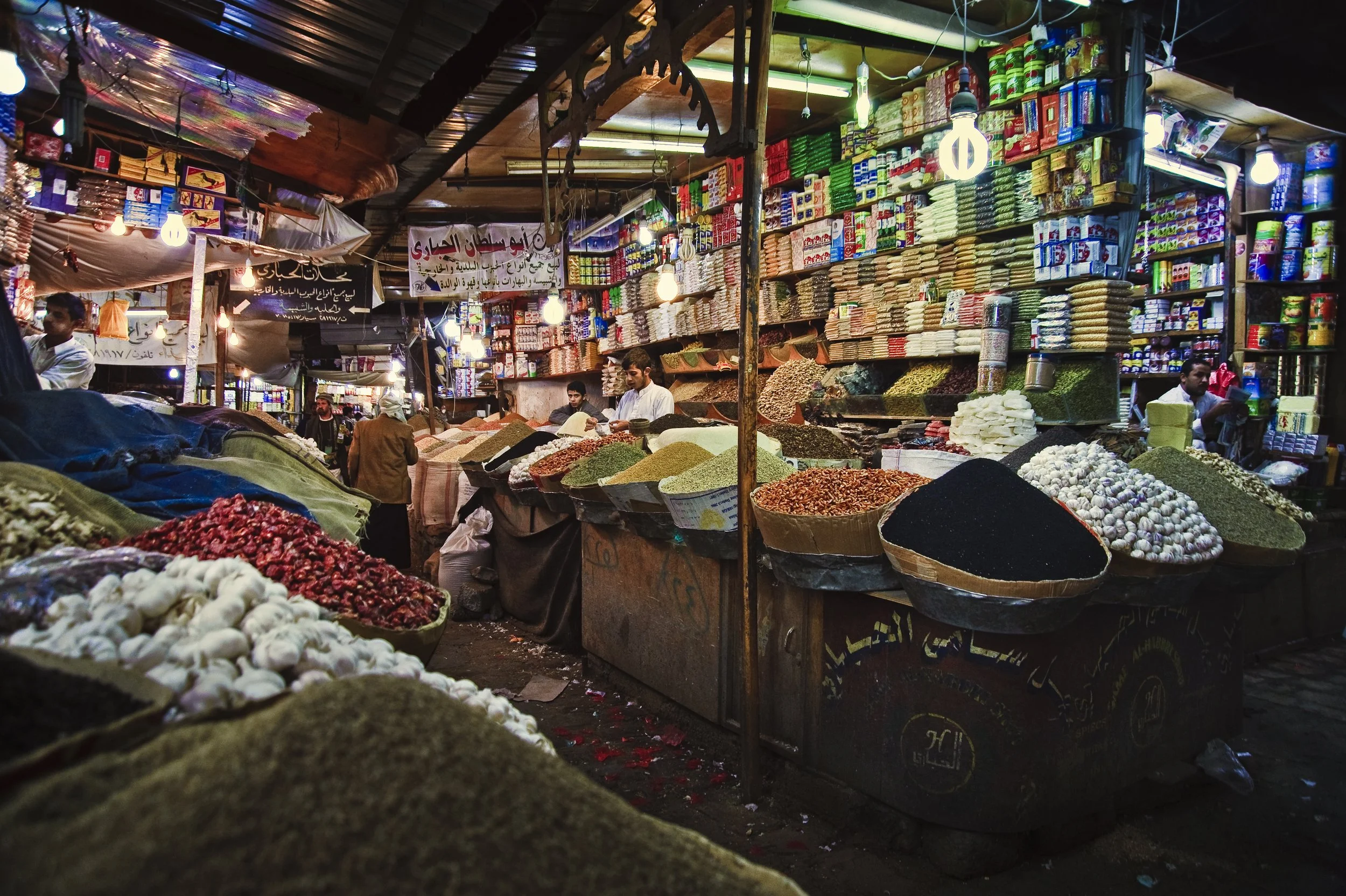 A bustling market stall with various spices and herbs in bowls, surrounded by colorful packaged goods, and shop vendors and customers in the background.