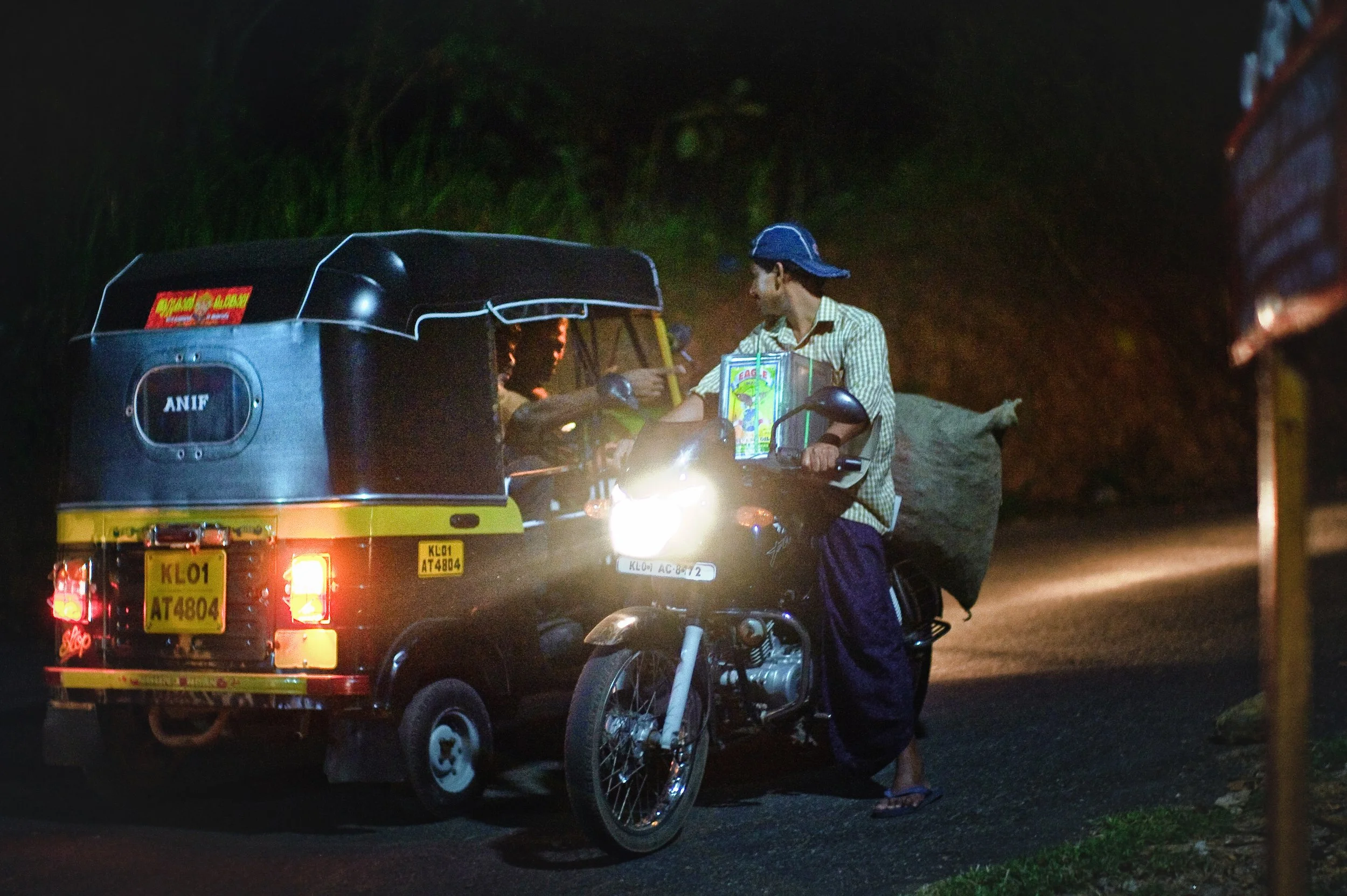 A man on a motorcycle with a large bag, waiting to board a black and yellow auto rickshaw at night. The motorcycle's headlight is on, illuminating the scene.