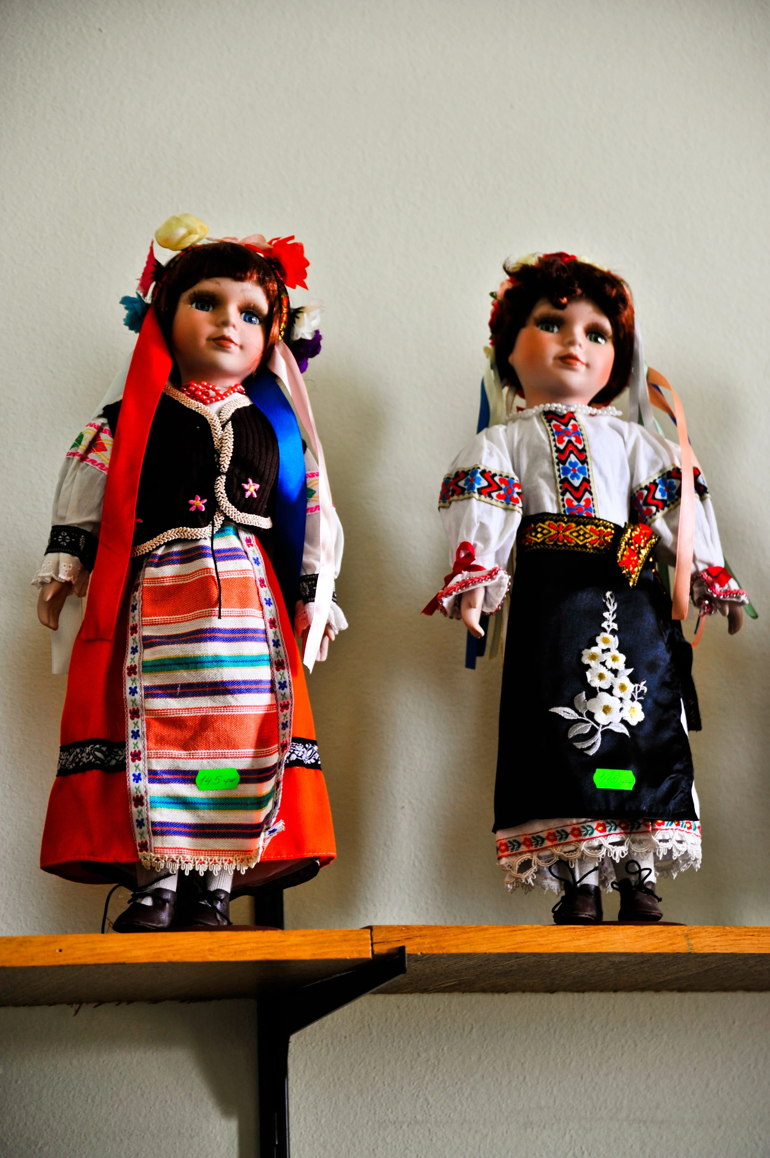 Two traditional dolls dressed in colorful folk costumes standing on a wooden shelf against a plain wall.