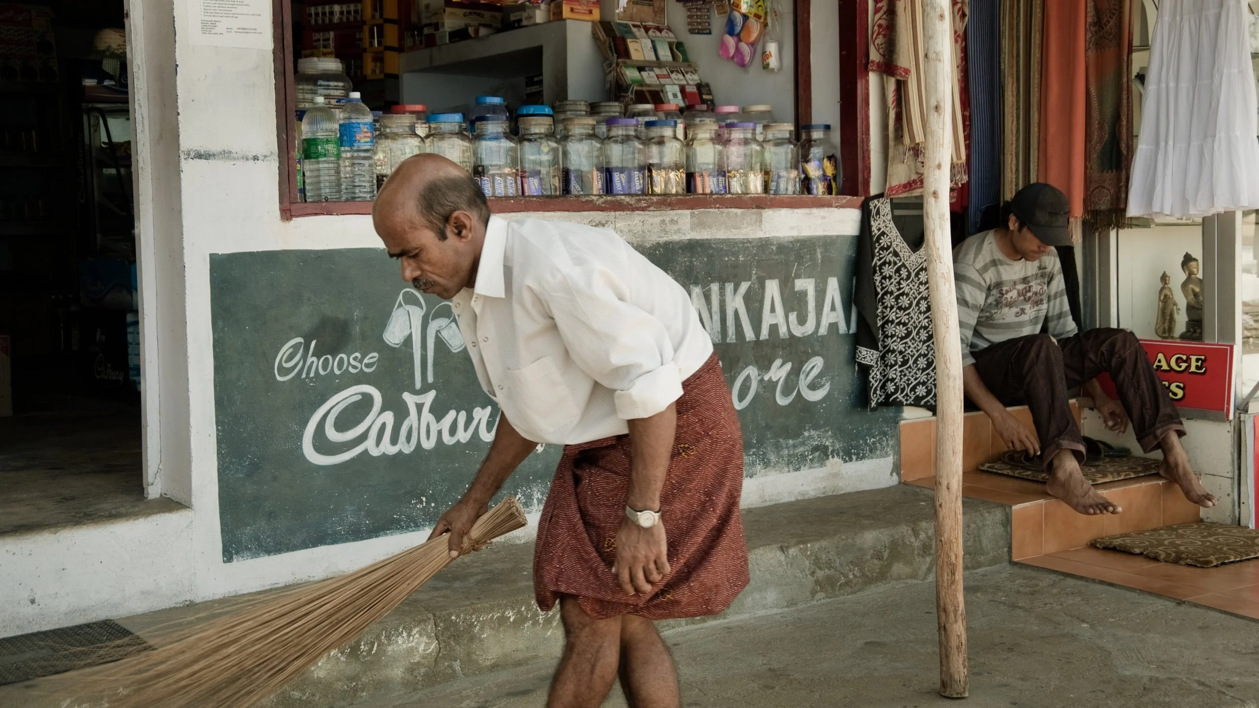 Man sweeping outside a shop with jars and textiles, another person sitting nearby