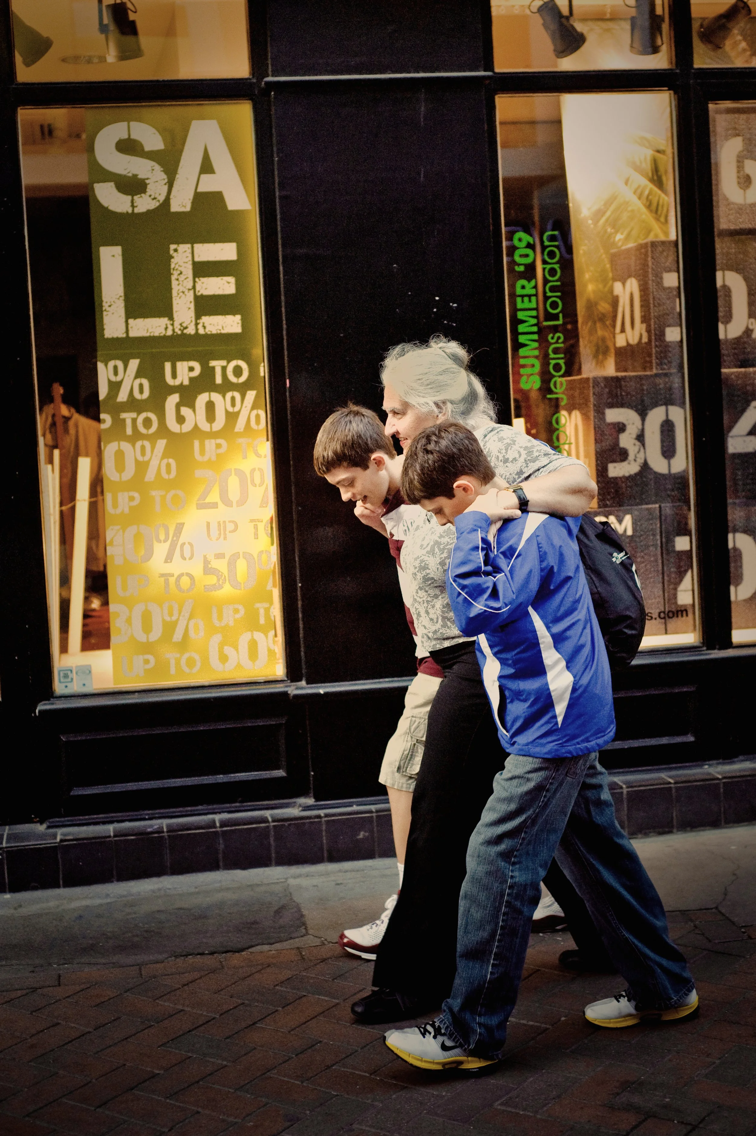 A woman walking on the sidewalk with two young boys in a city in front of a store window with sale signs.