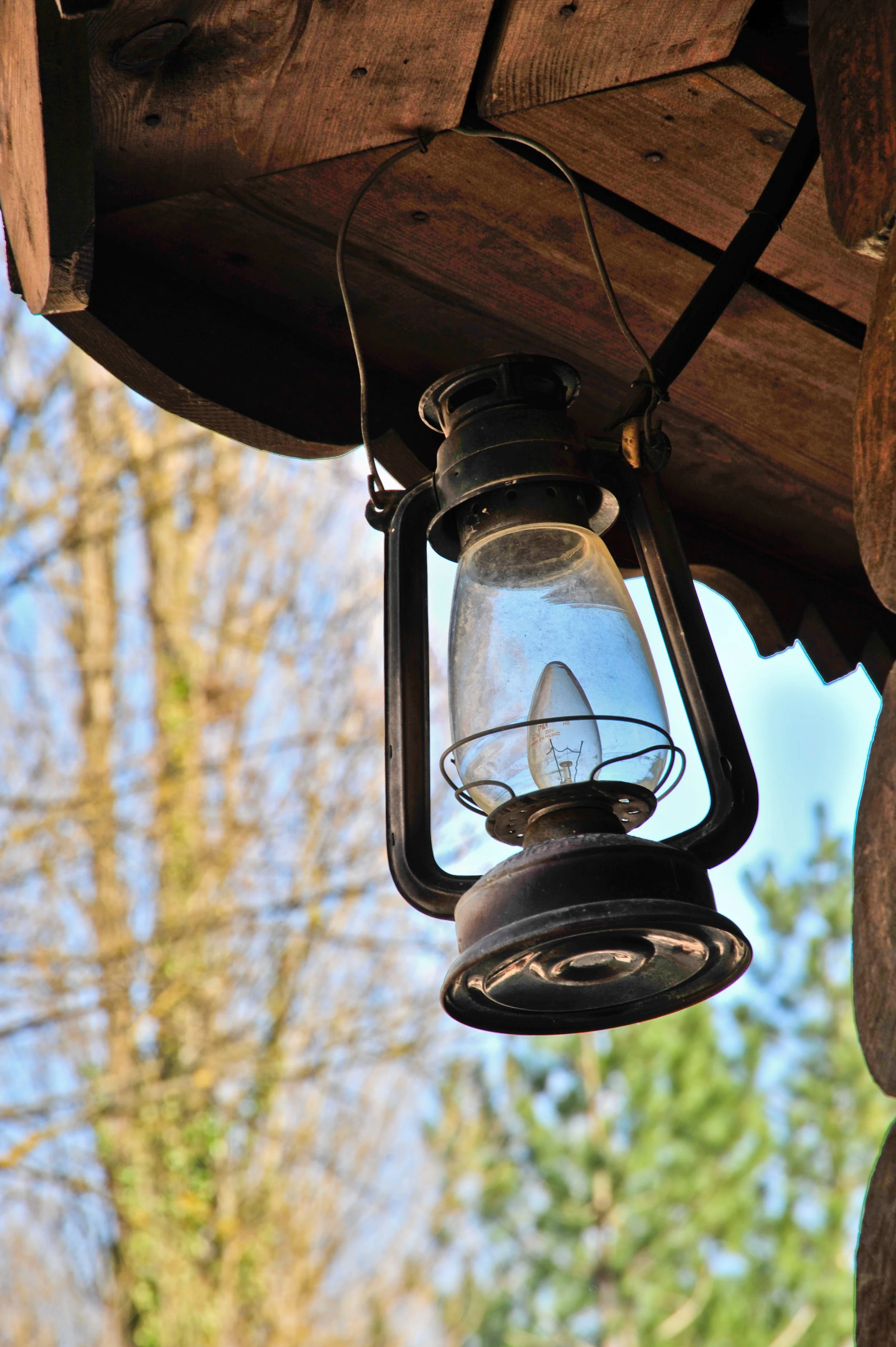 An antique hanging lantern with a glass enclosure, mounted on the corner of a wooden structure outdoors, with trees and blue sky in the background.