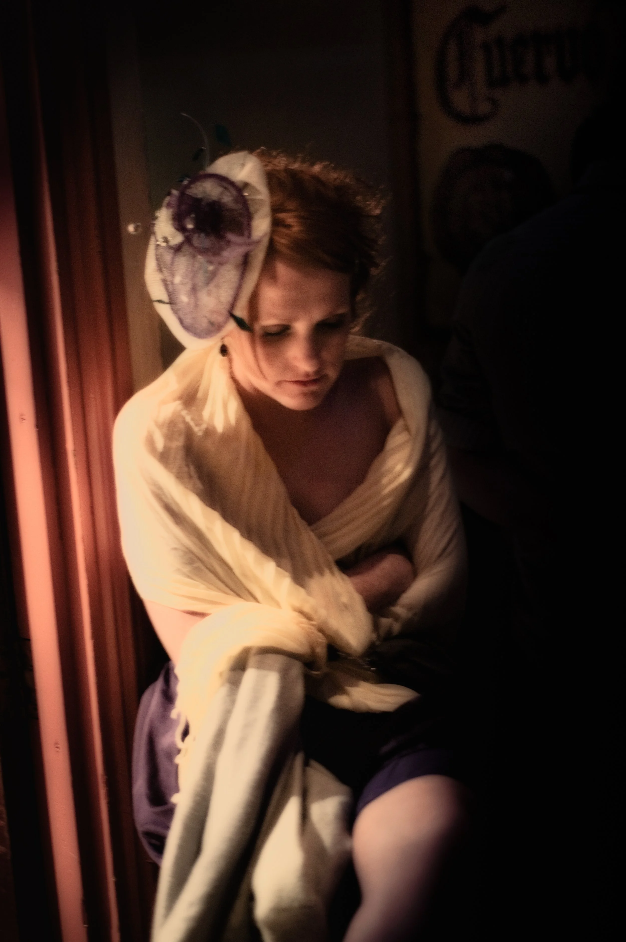 A woman with short, curly hair wearing a fancy hat with purple accents, wrapped in a yellowish shawl, sitting in a dimly lit environment, looking down.
