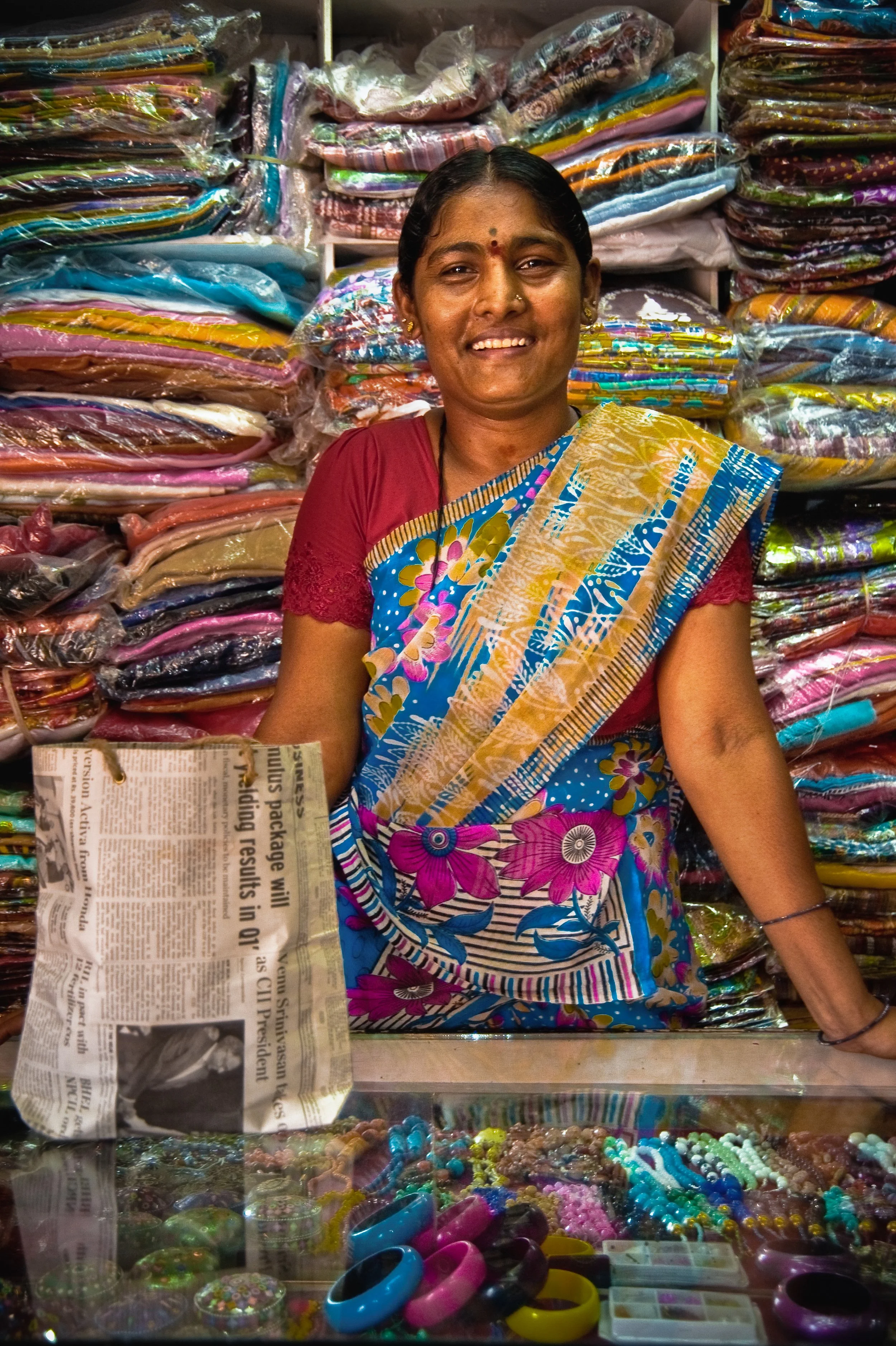 Smiling woman wearing a colorful sari, standing behind a display case with jewelry, in a shop filled with neatly folded clothes on shelves in the background.