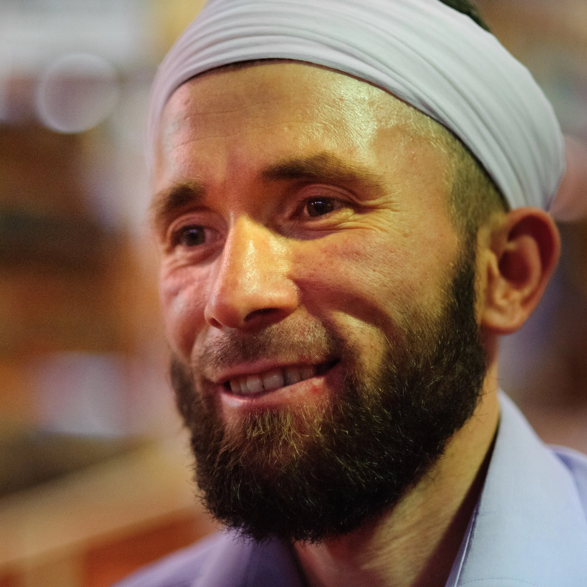 Close-up of a smiling man wearing a white head covering and a light blue shirt, with a blurred background.