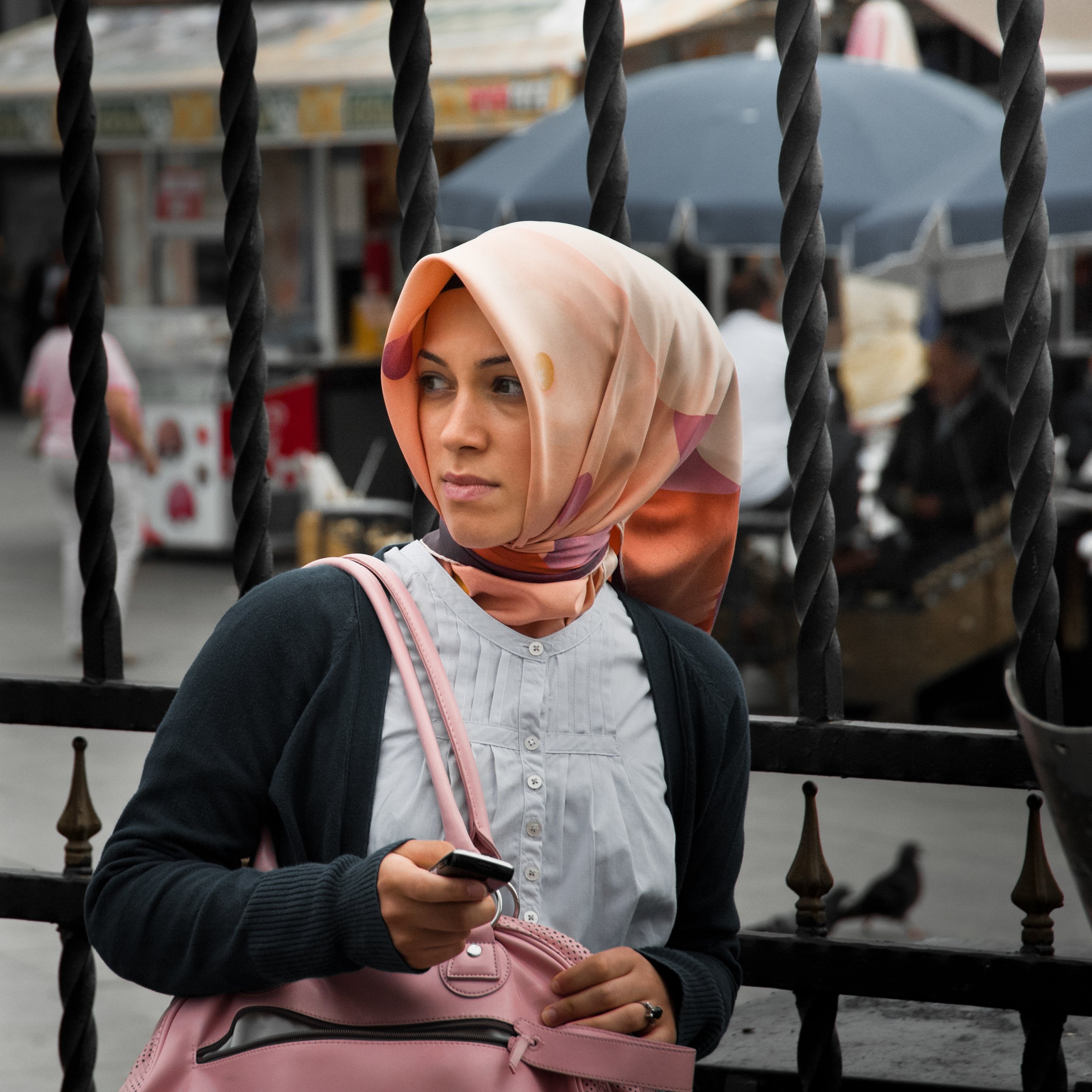 Woman with a peach-colored patterned headscarf holding a smartphone and a pink bag, standing behind a black iron fence in an outdoor market.