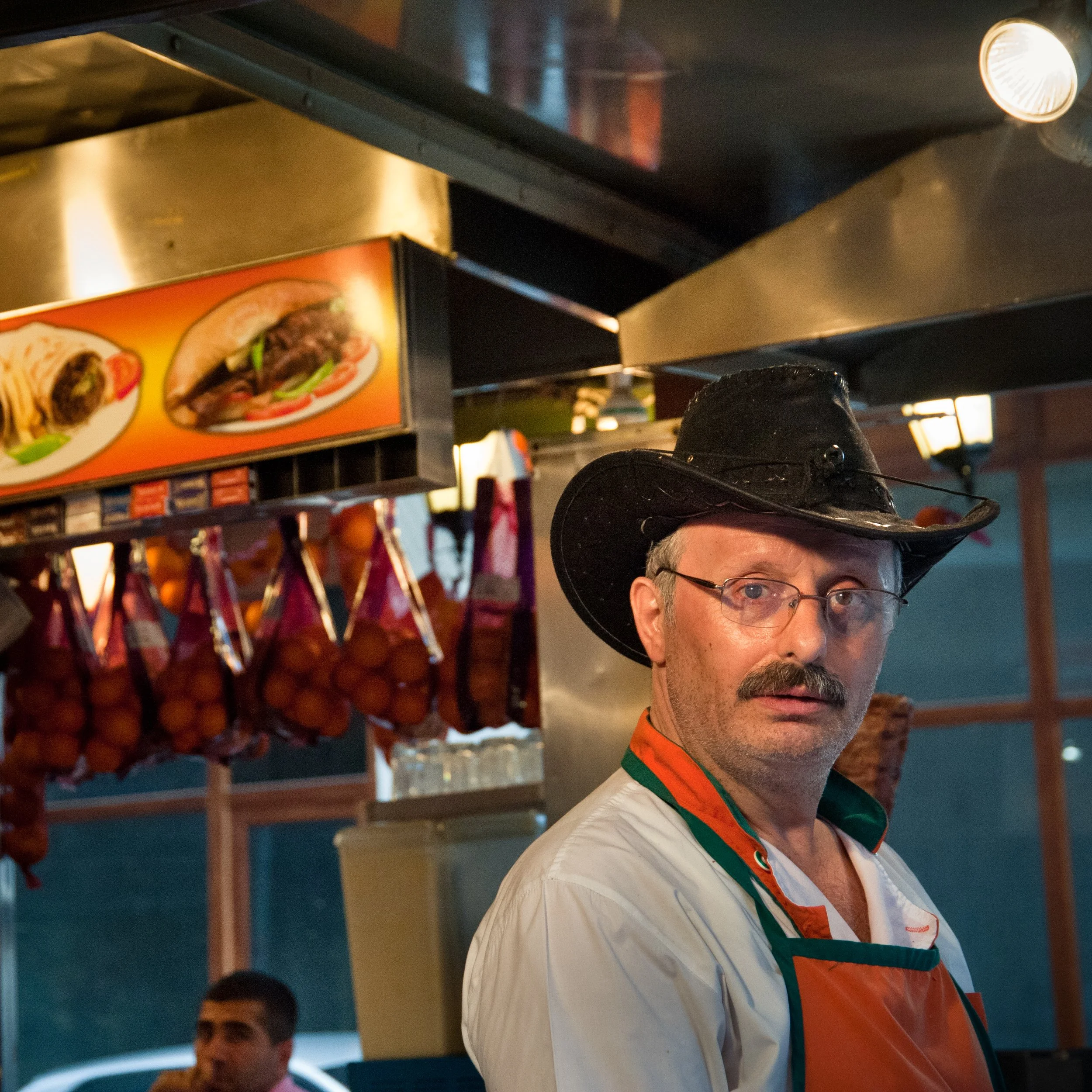 Man wearing a black cowboy hat, glasses, and a white apron with orange and green trim inside a food stand, with hanging sausages and a menu with images of hot dogs or sandwiches.