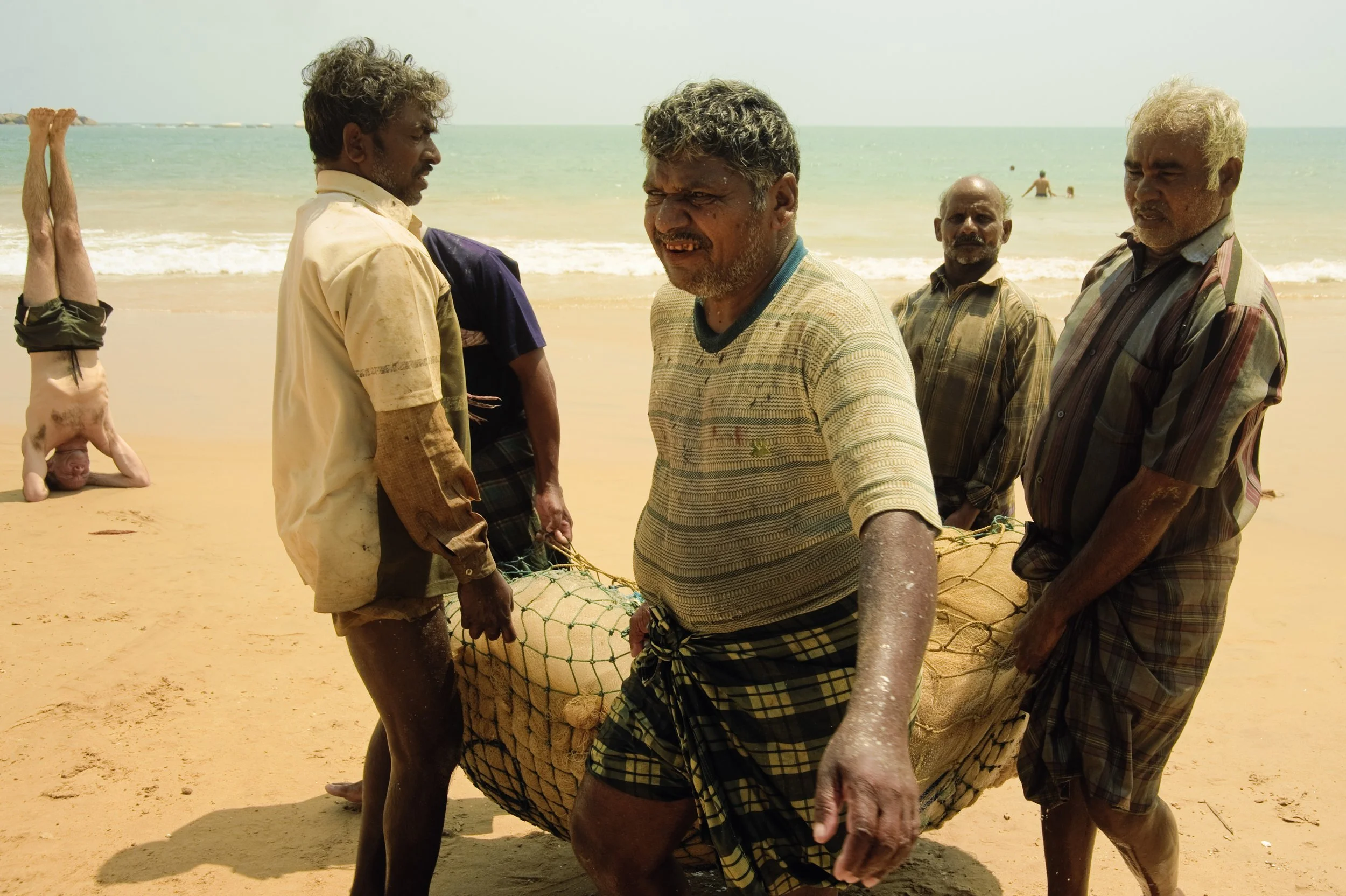 Five men carrying a fishing net on a beach with people swimming in the ocean in the background.