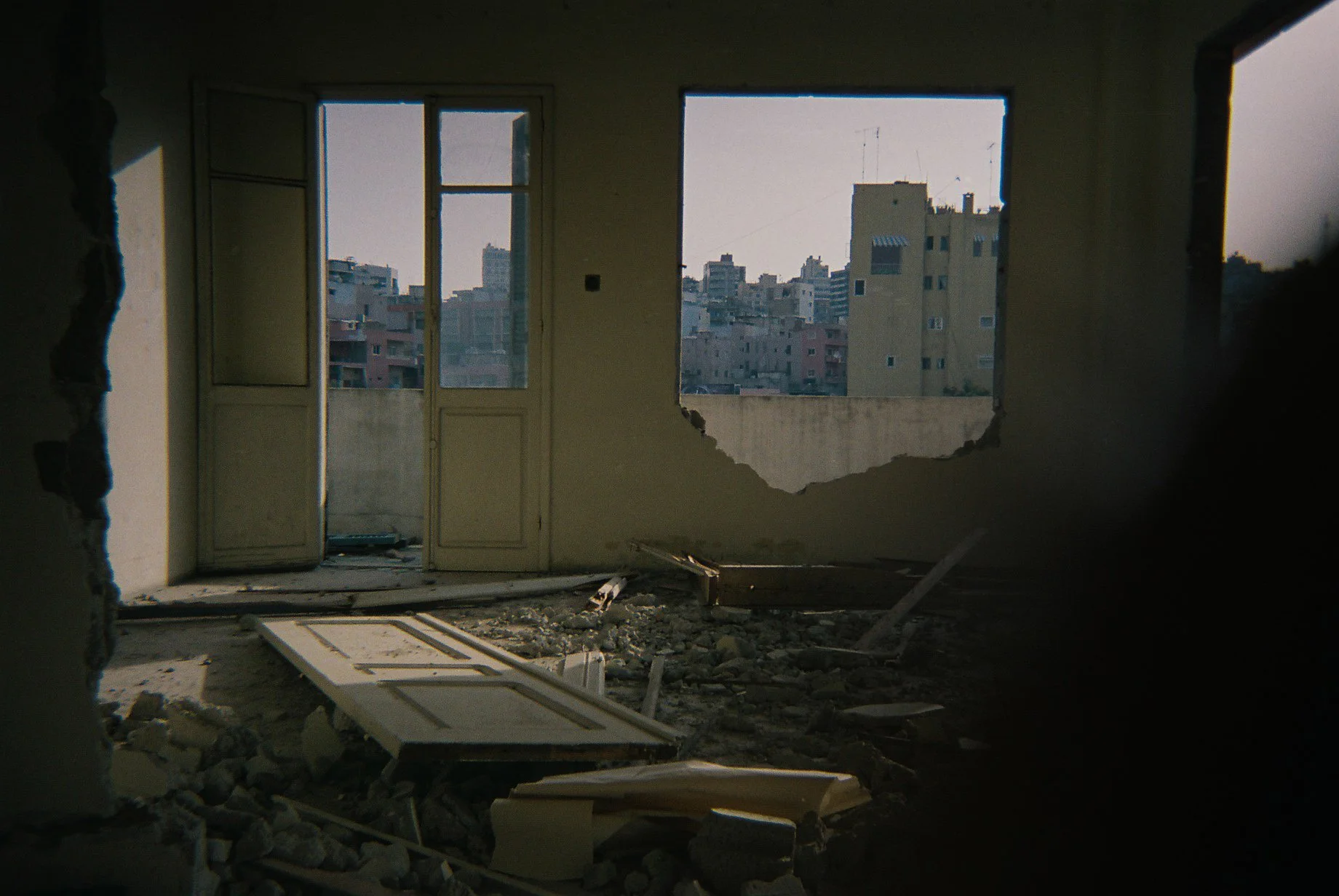 Interior of a damaged building with debris on the floor, a broken door, and large windows showing a cityscape with multiple buildings outside.