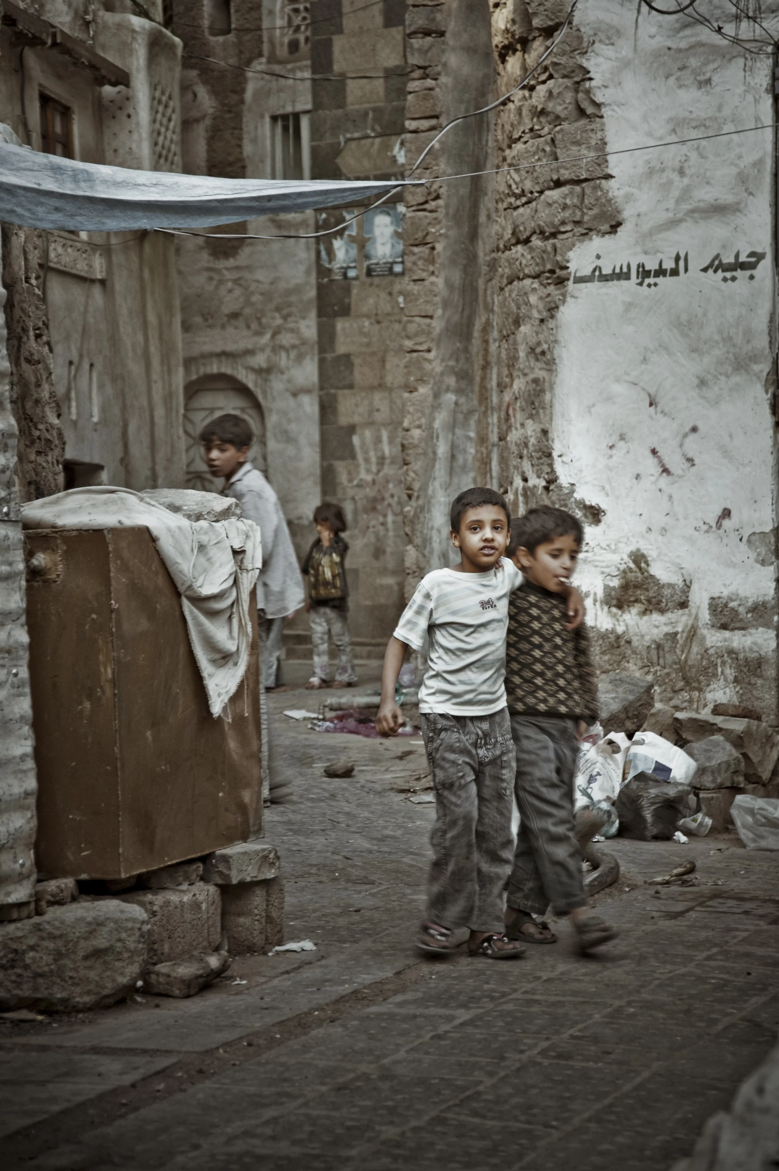 A group of children playing and walking in a narrow, rustic alleyway with stone walls and scattered trash.