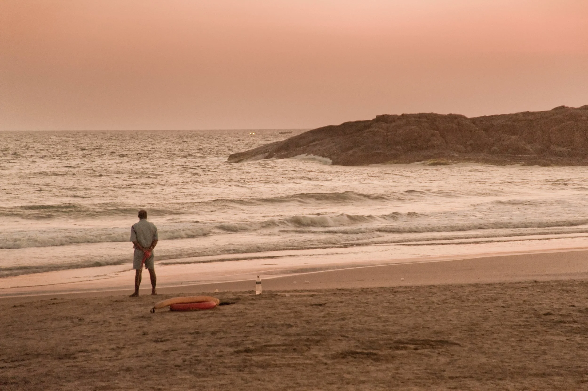 A man stands on a beach facing the ocean, with a life preserver and water bottle on the sand nearby, during sunset or sunrise.