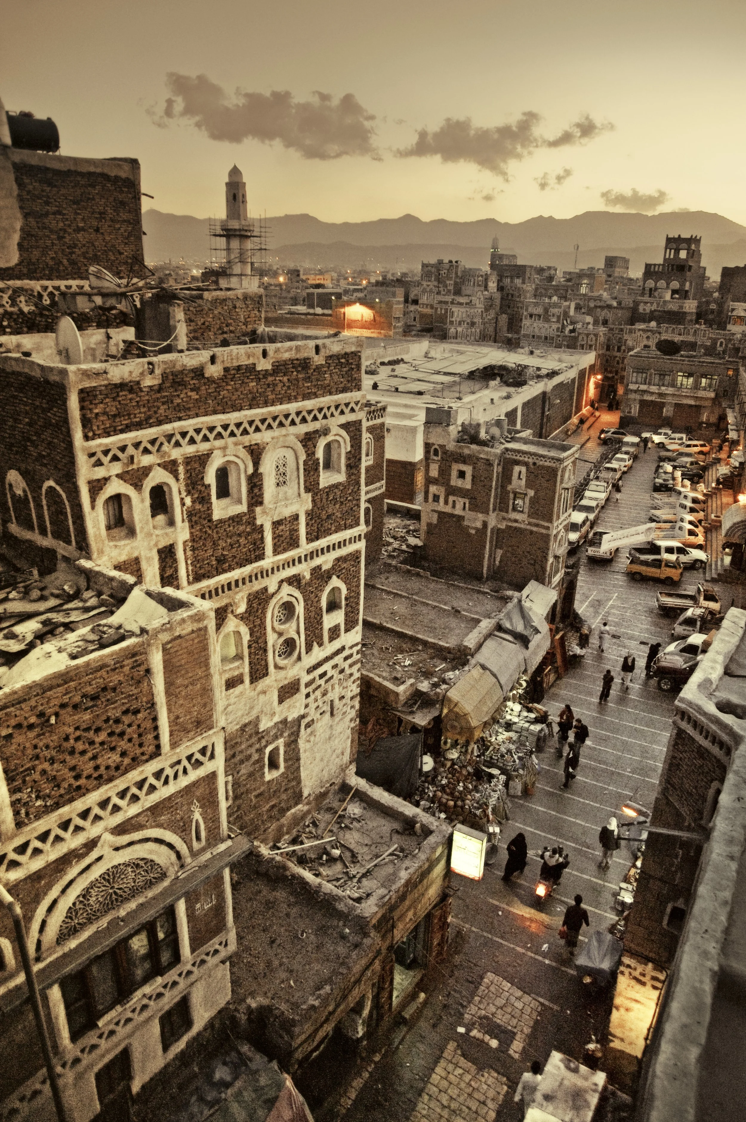 Cityscape at sunset with historic brick buildings, parked cars, and pedestrians on a busy street.