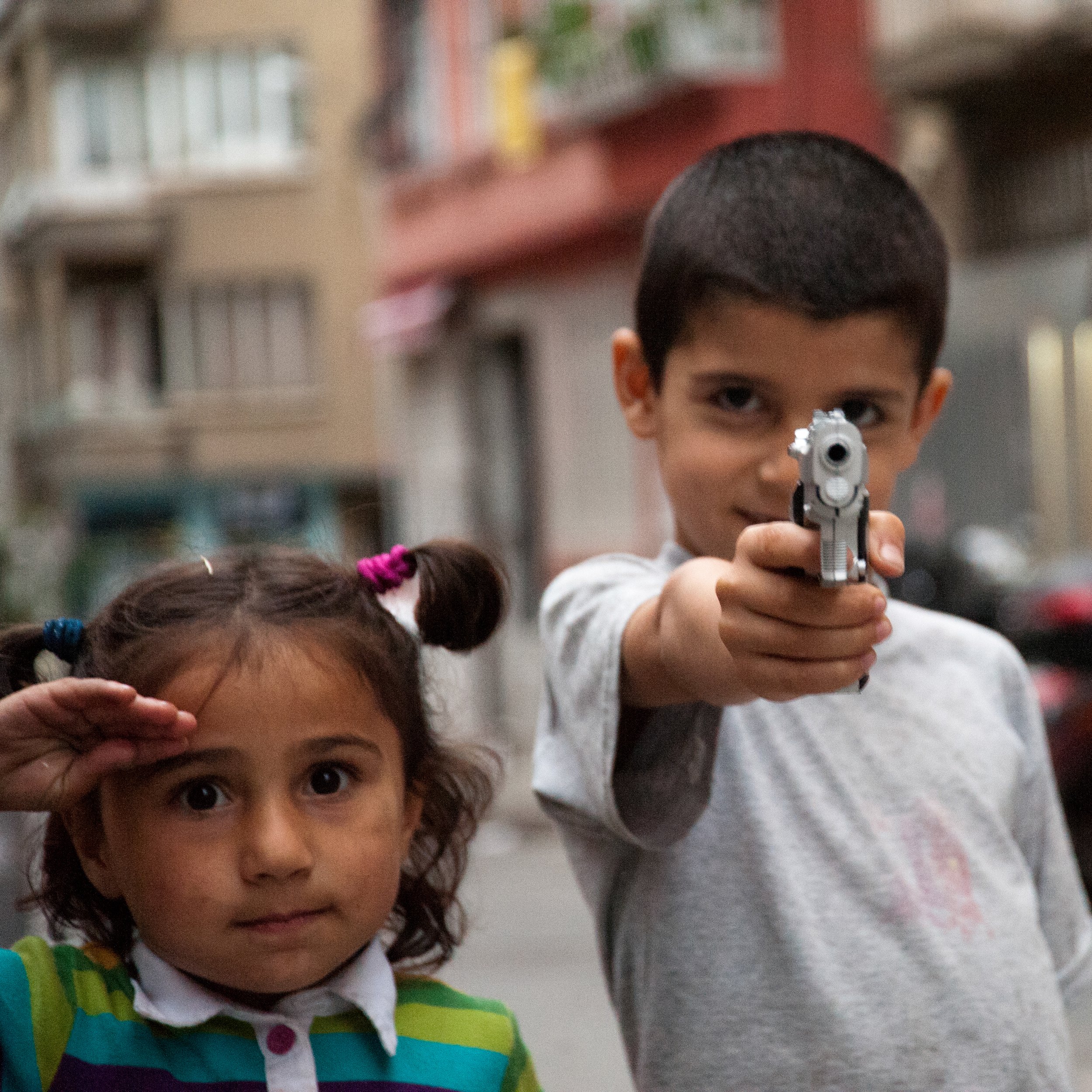 A young boy aiming a toy gun at the camera and a girl saluting with her hand, on a city street with apartment buildings in the background.