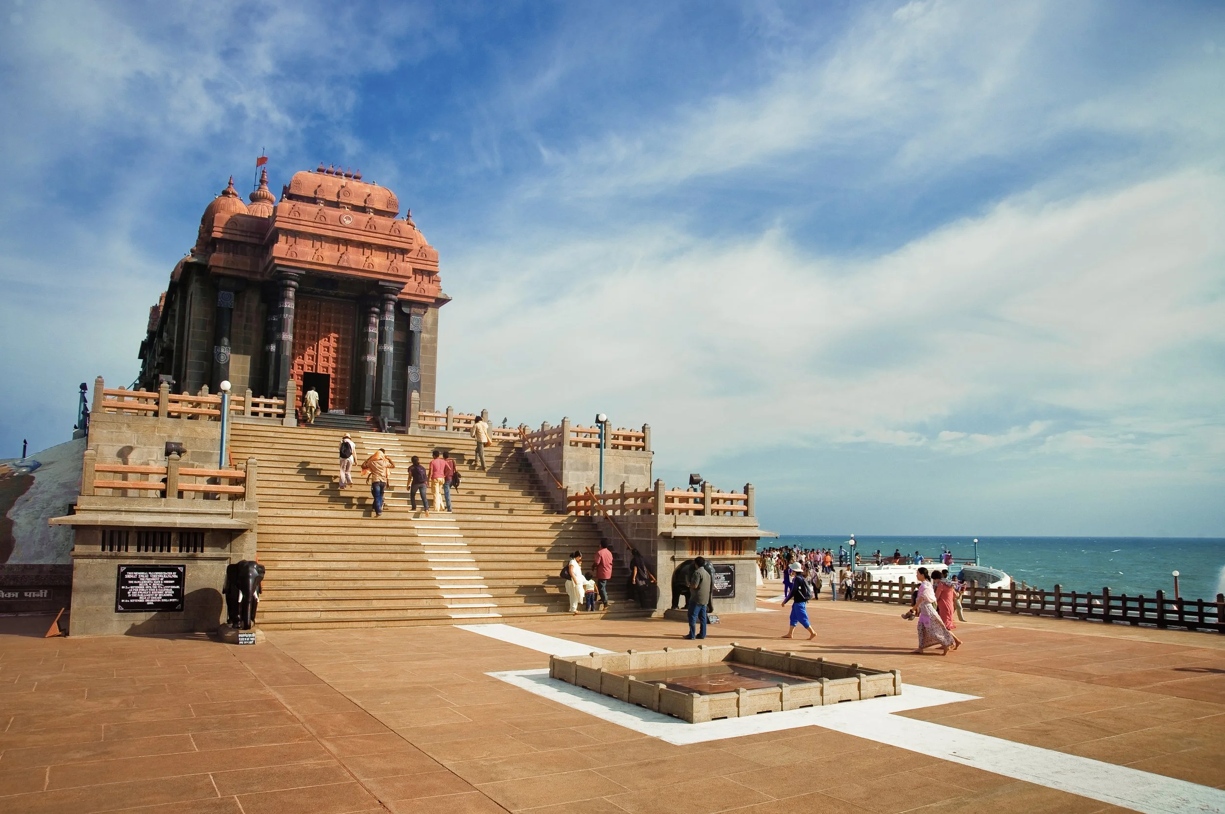 A temple-like structure with red stone architecture situated near the coast, with stairs leading up to the entrance and people walking around the area against a blue sky and ocean background.