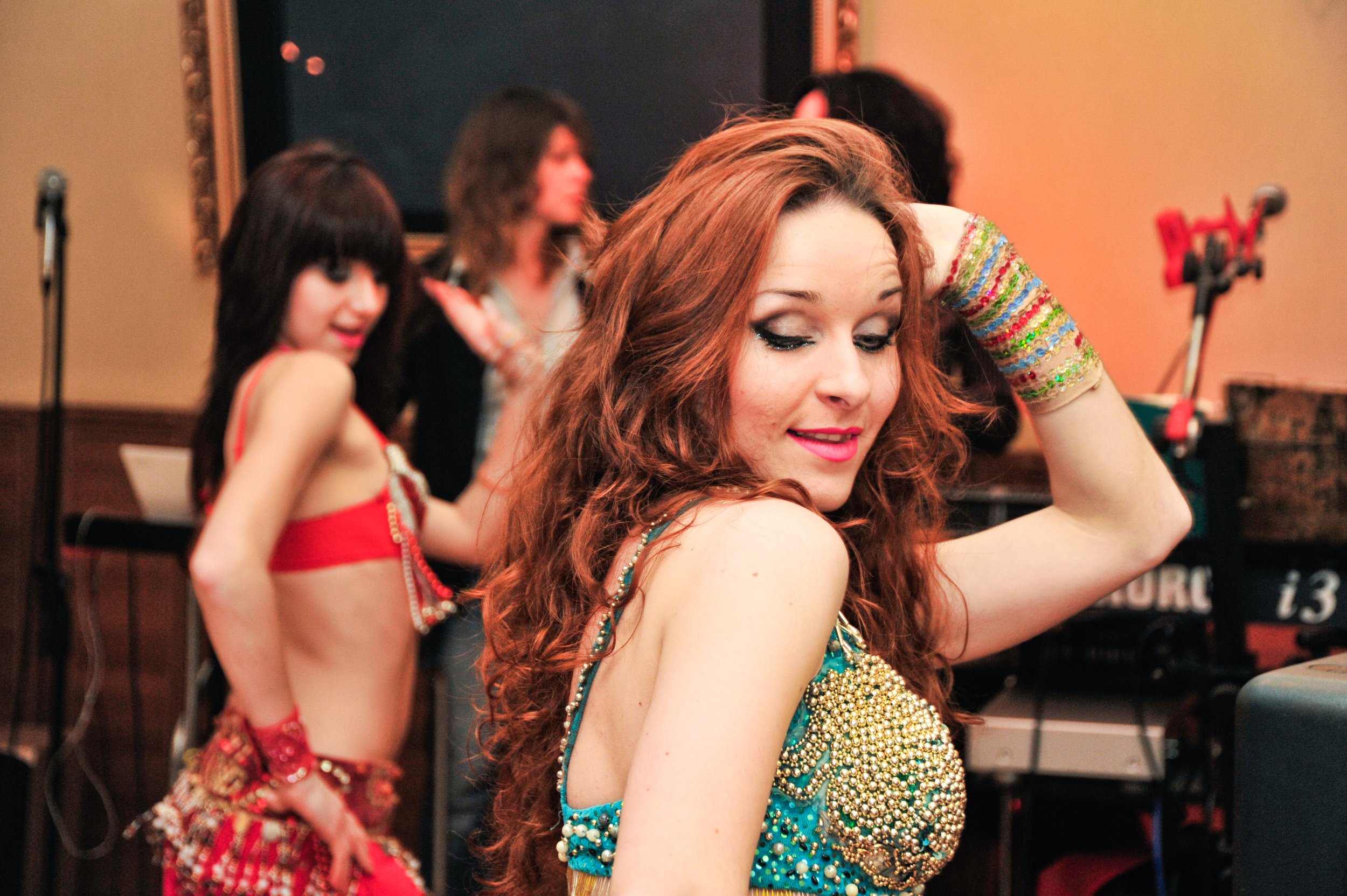 Red-haired woman dancing with colorful jewelry and a beaded top, with other women dancing in the background at a party.
