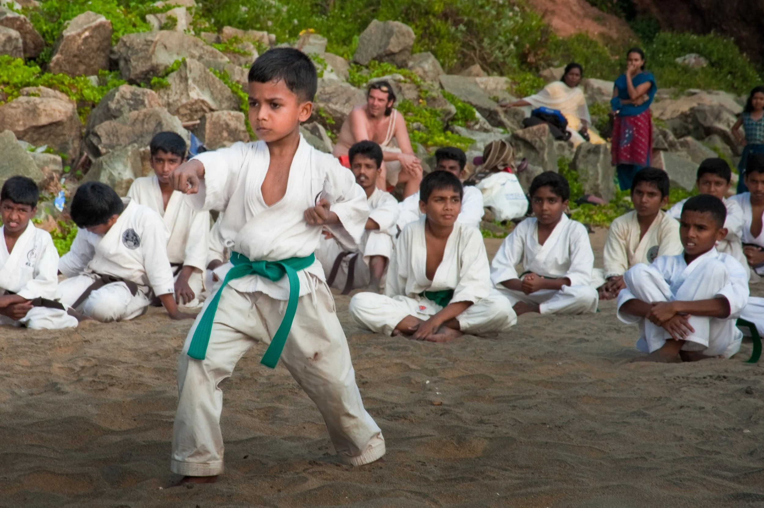 Young boy practicing martial arts or karate on a sandy outdoor area with other children and adults sitting and standing nearby, surrounded by rocks and greenery.