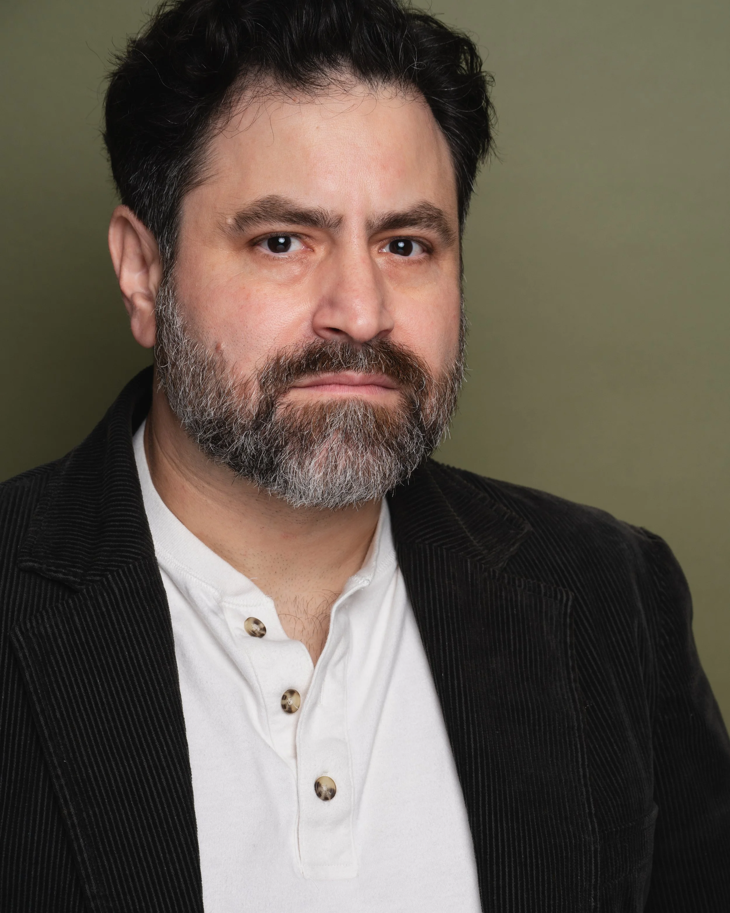 Close-up portrait of a middle-aged man with dark hair, a beard, and mustache, wearing a white collared shirt and a black jacket, against a plain olive-green background.