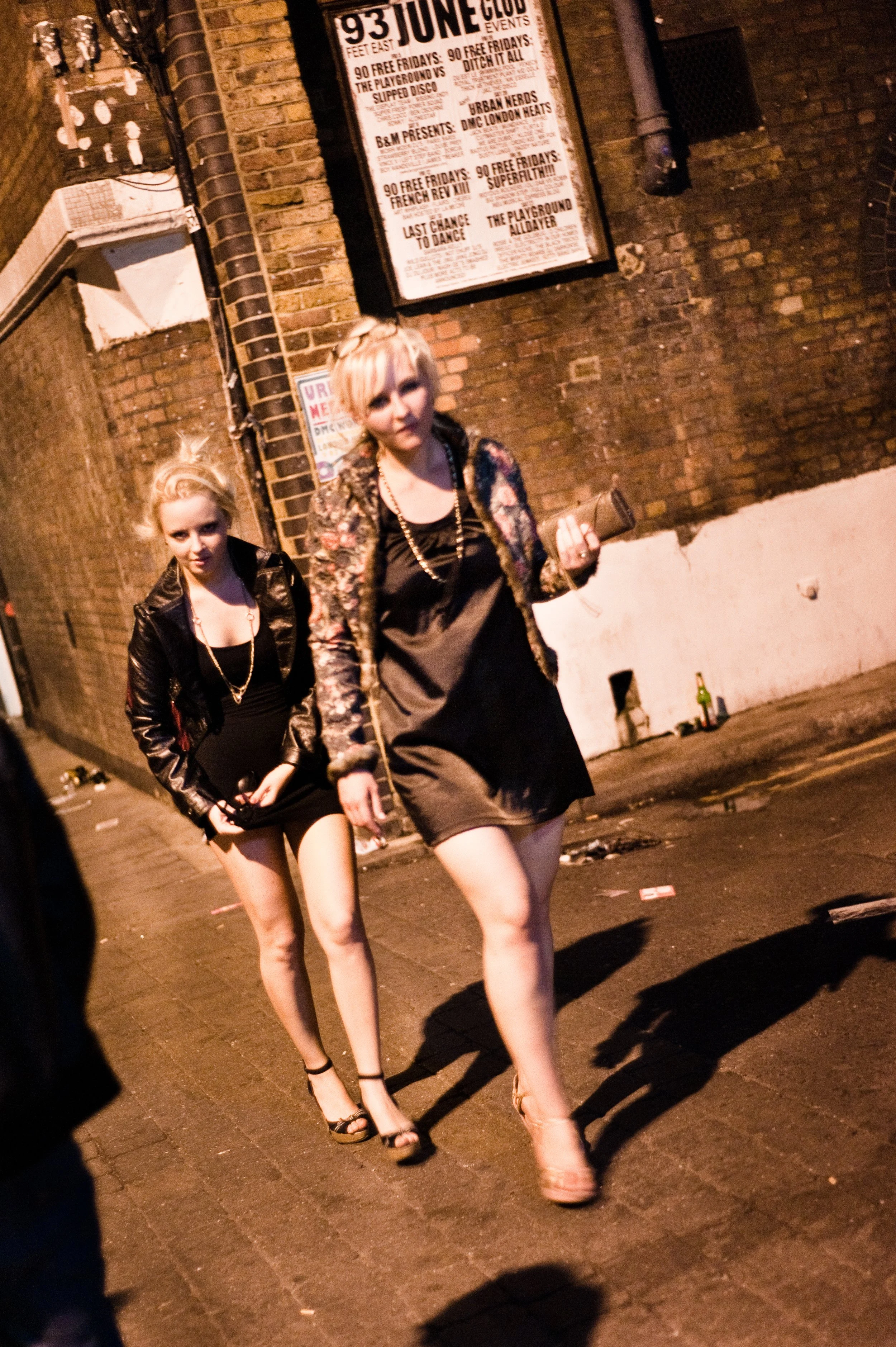 Two young women walking on a city sidewalk at night. They are dressed in dark clothing with high heels. One woman is in front, wearing a floral jacket and holding a clutch, the other behind, wearing a black leather jacket and a short dress. There is 