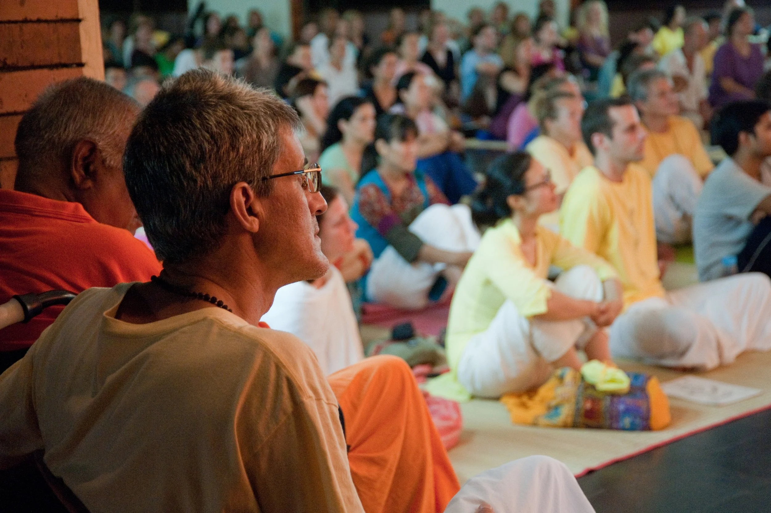 A diverse group of people sitting cross-legged on the floor, attentively listening during a meditation or spiritual gathering, with some wearing traditional Indian attire.