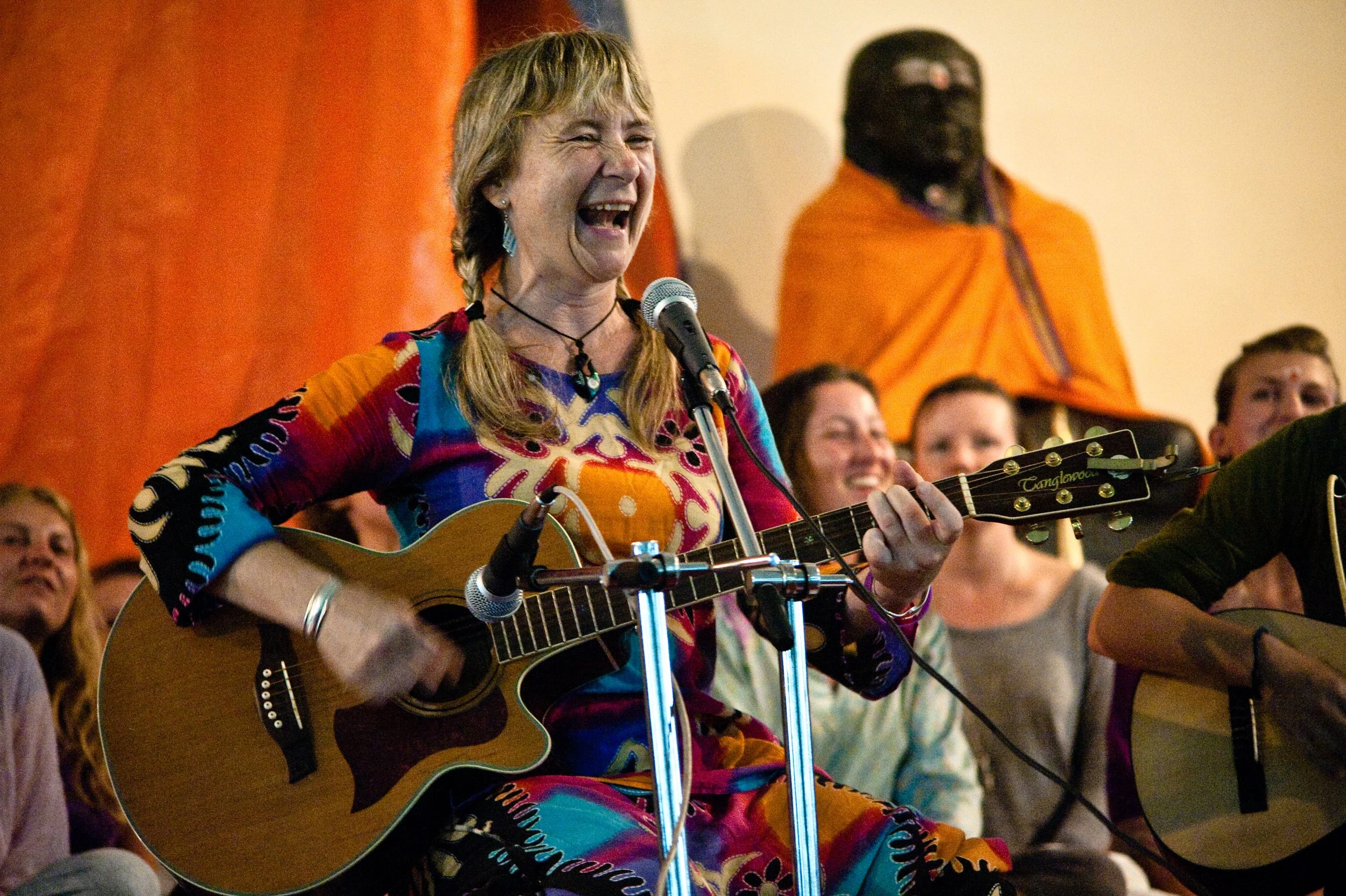 A woman with blonde hair in pigtails is playing a guitar and singing into a microphone, smiling happily while sitting among a group of people. The background includes a person in an orange robe and an orange curtain.