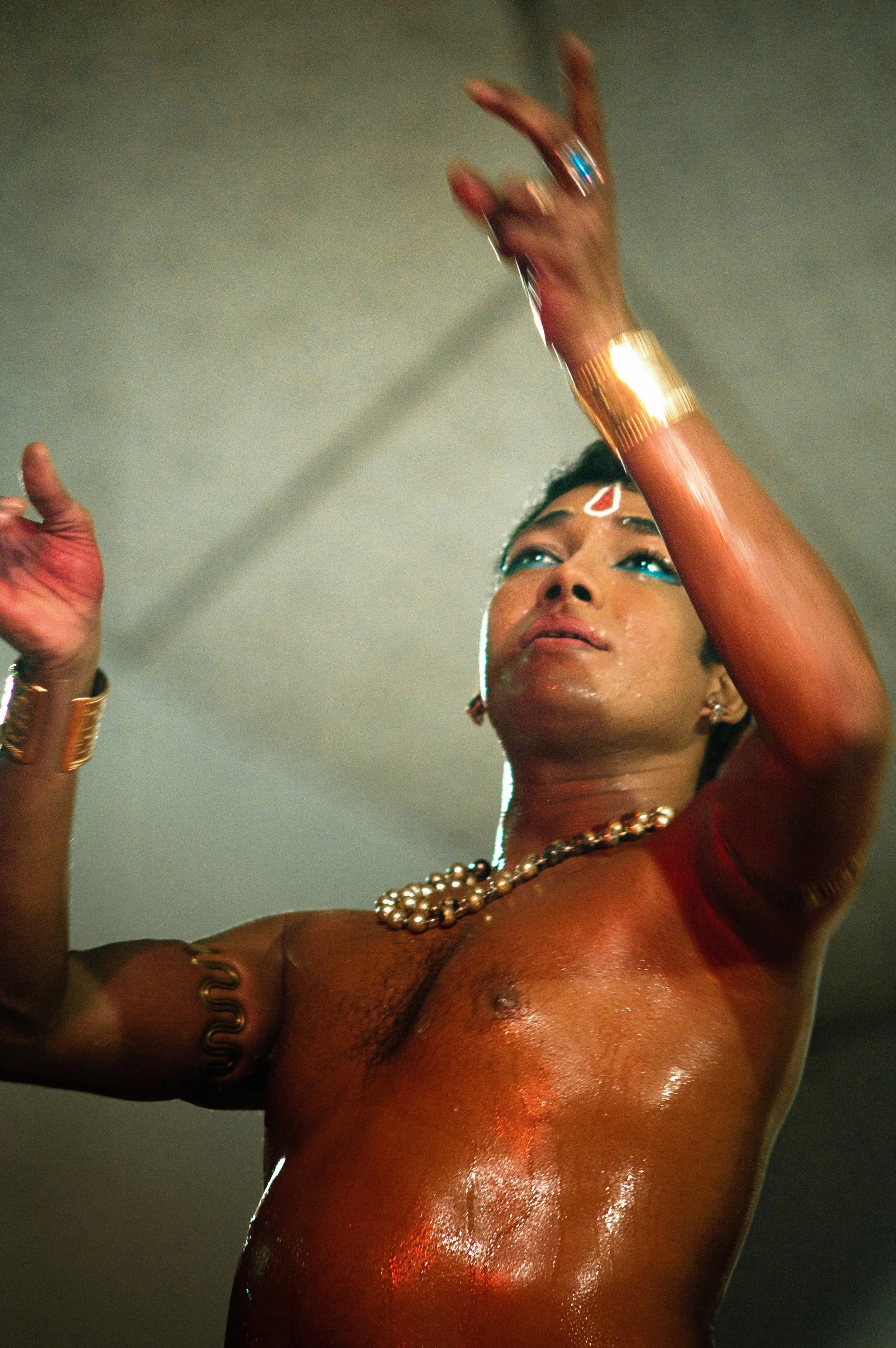 A person with traditional Indian jewelry on their arms and neck, wearing blue eye makeup and a red mark on their forehead, is raising their hands in a dance gesture. Their chest is glistening with sweat.